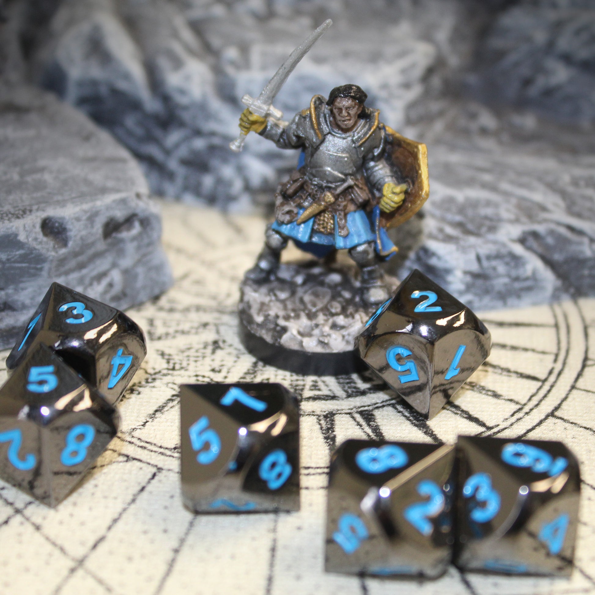 Metal Black Frost dice set scattered on a tan play mat with a mini figure and rocky set pieces behind them