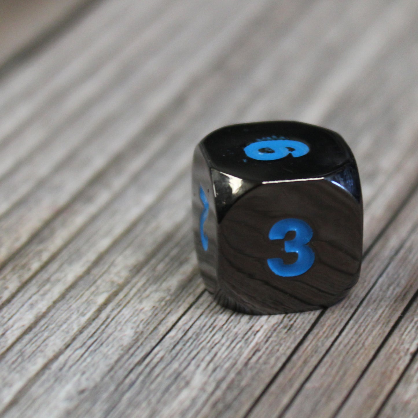 Black Frost D6 die on a wooden surface. The close-up of the die showcases the shiny black surface and the blue numbering.