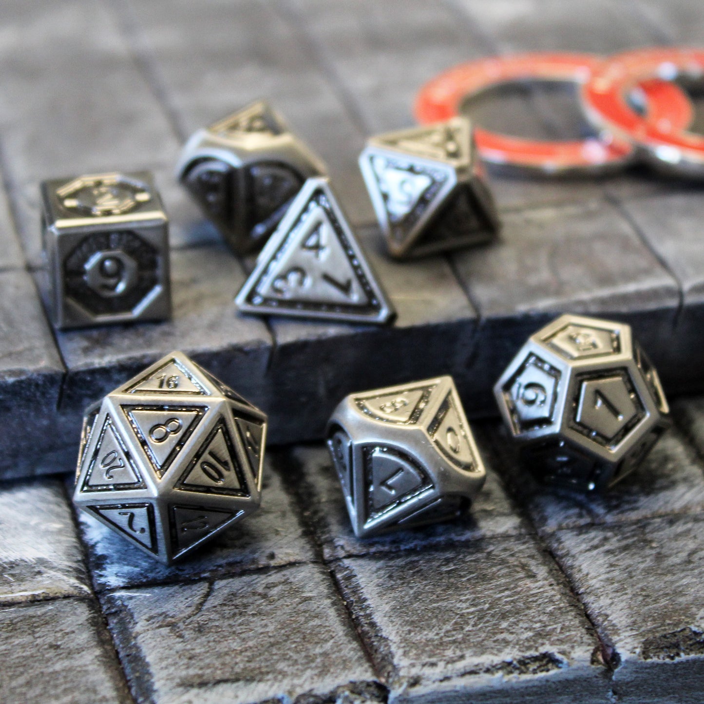 Metal Bullseye dice set arranged on top of a gray stone floor set piece. Two red condition rings are out of focus in the background.
