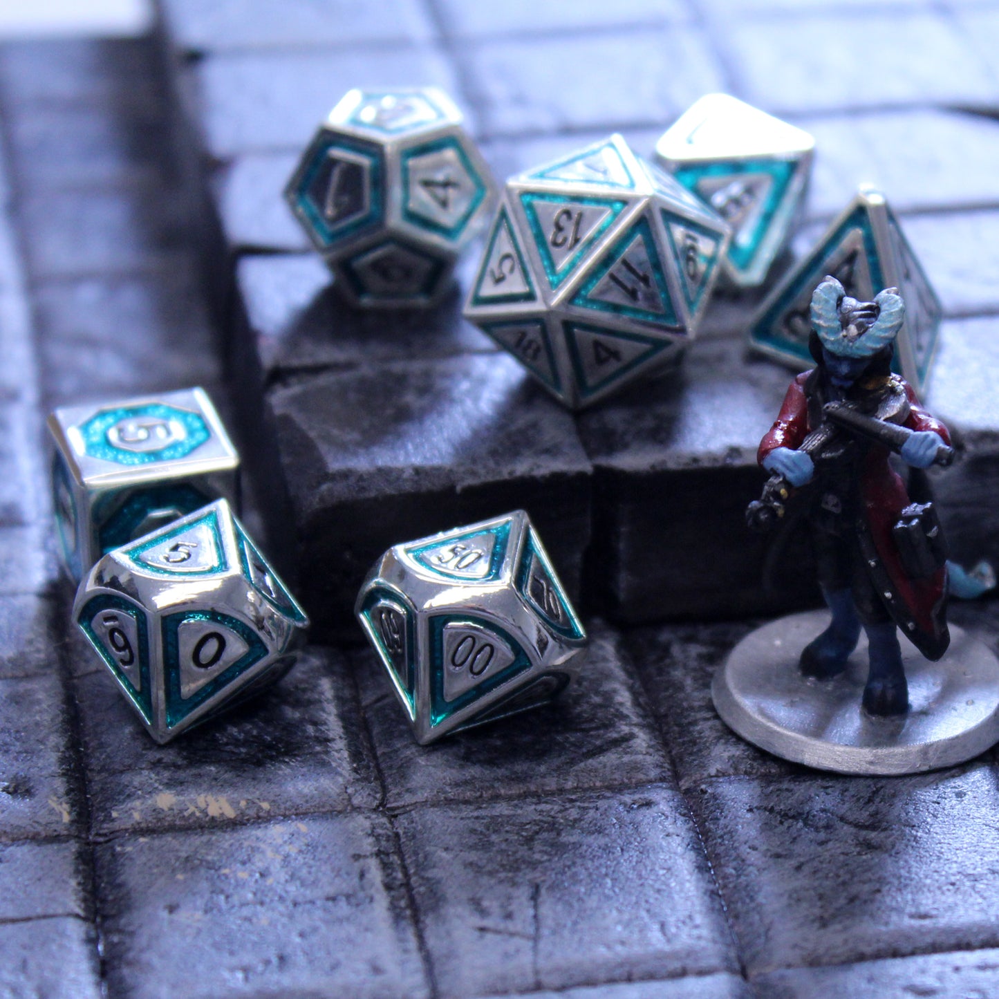 Closeup photograph of silver dice with teal inlays on a grid next to a small figure of a horned being.