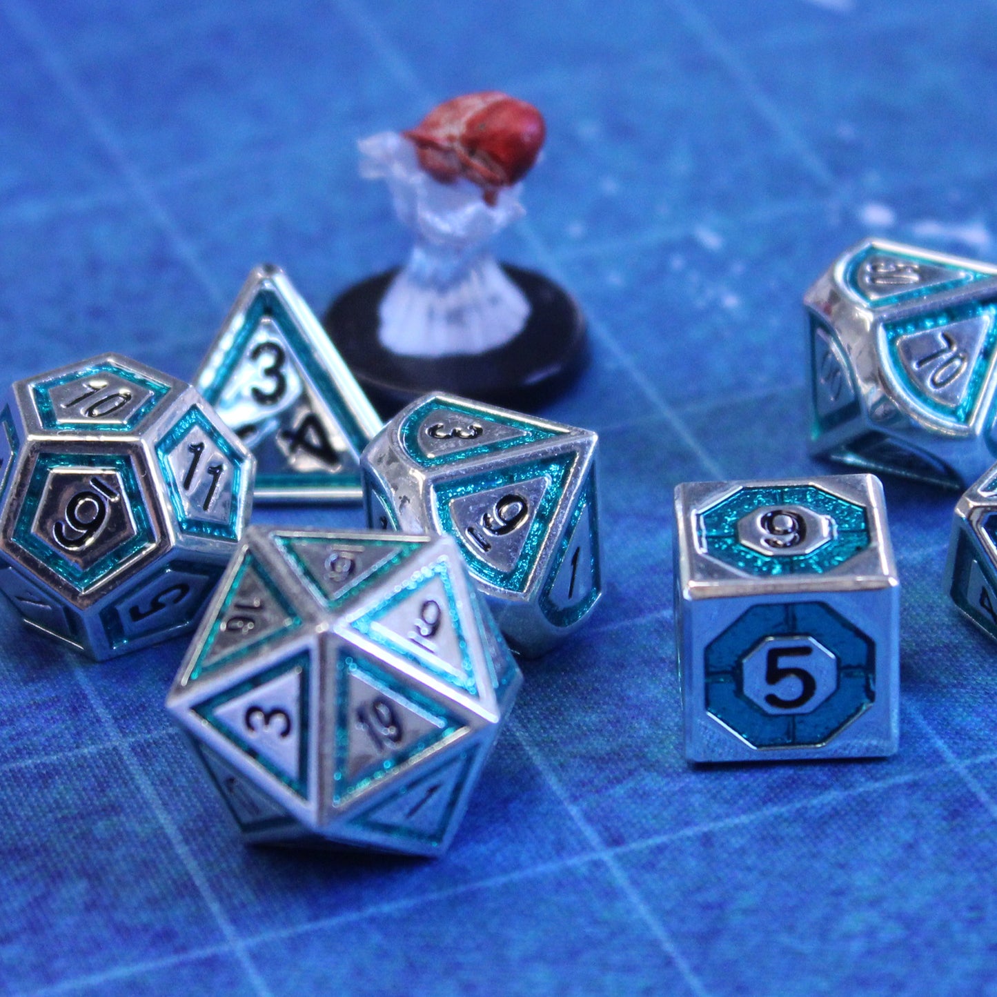 Closeup photo of silver dice with a bullseye-shaped teal inlay on blue gaming mat with a figure of a fish nearby.
