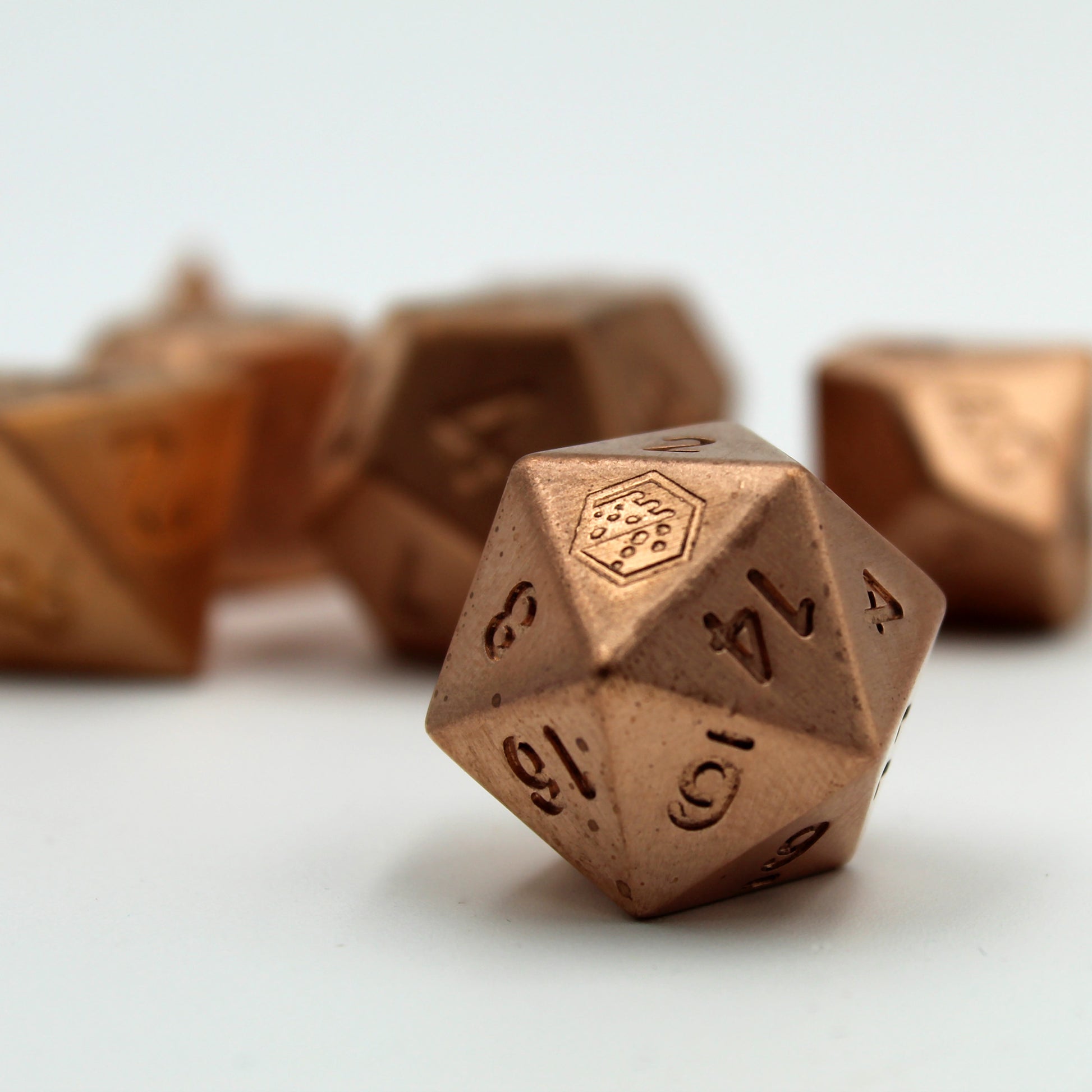 A close up of the D20 metal copper die on a white surface, showing the Dice Dungeons logo in place of the 20 and the 14 side facing forward. Other dice in the set are arranged in the background, but out of focus.
