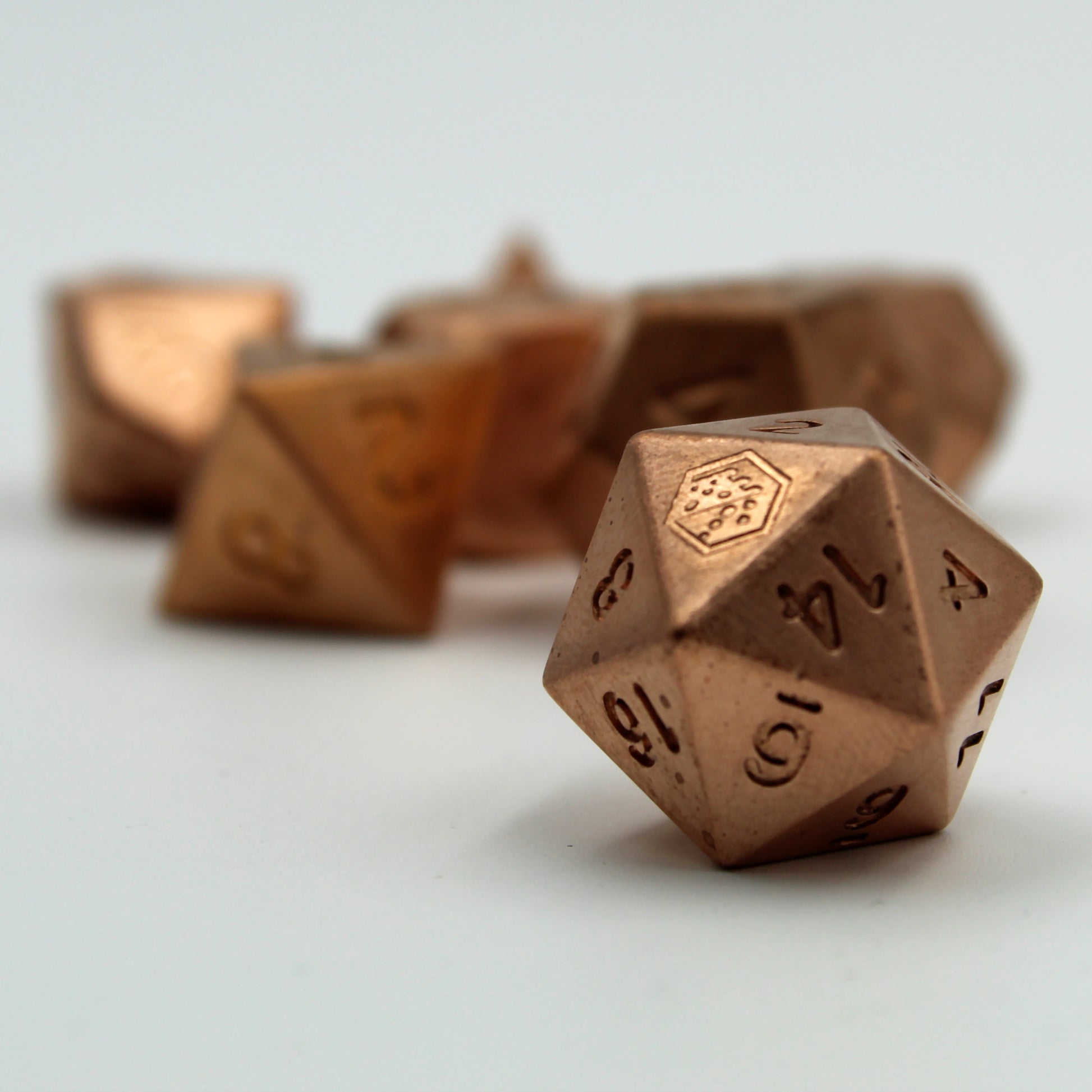 A close up of the D20 metal copper die on a white surface, showing the Dice Dungeons logo in place of the 20 and the 14 side facing forward. Other dice in the set are arranged in the background, but out of focus.