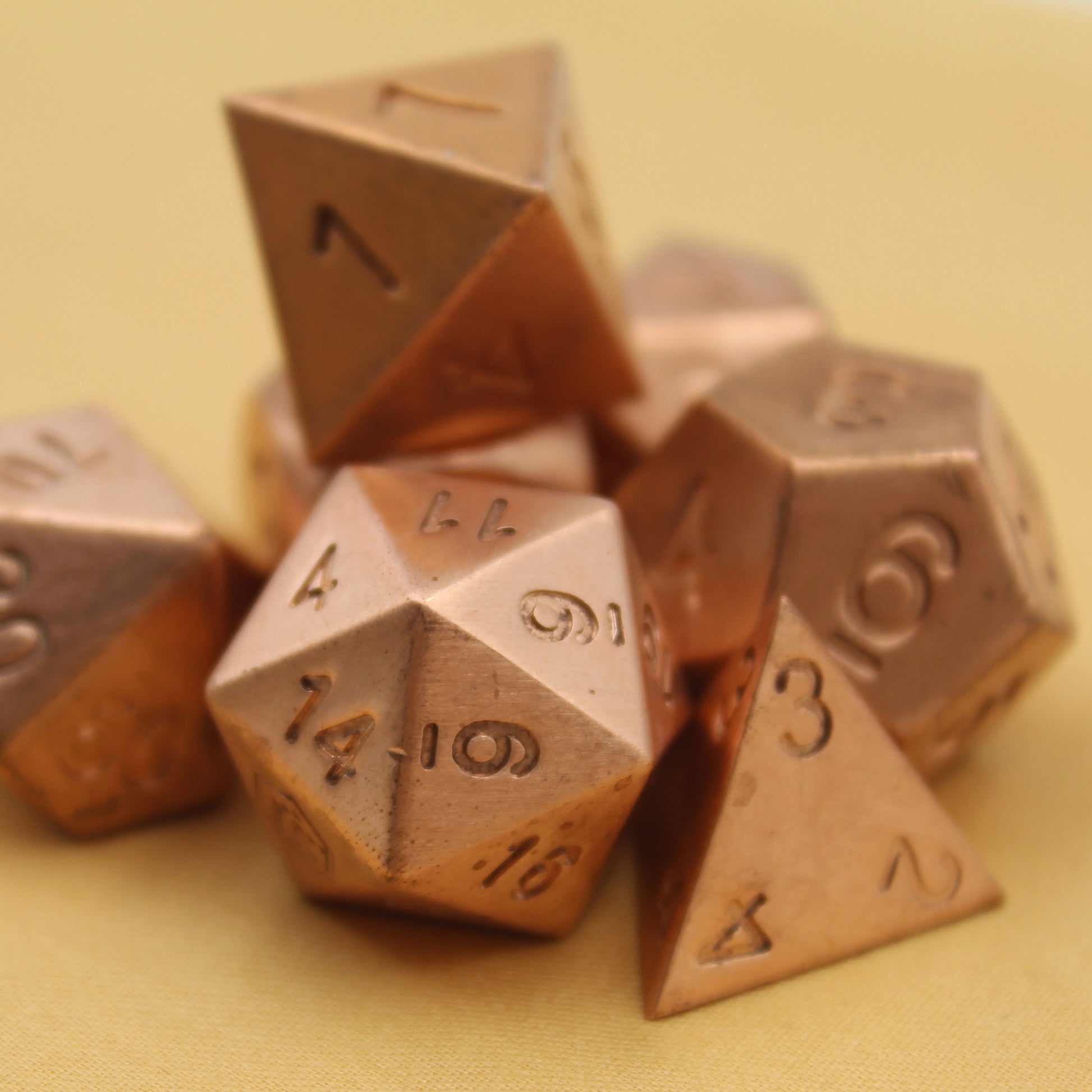 The metal copper dice arranged in a pile on a tan surface. The D20 die is in front, with the 14 and 6 sides facing forward.