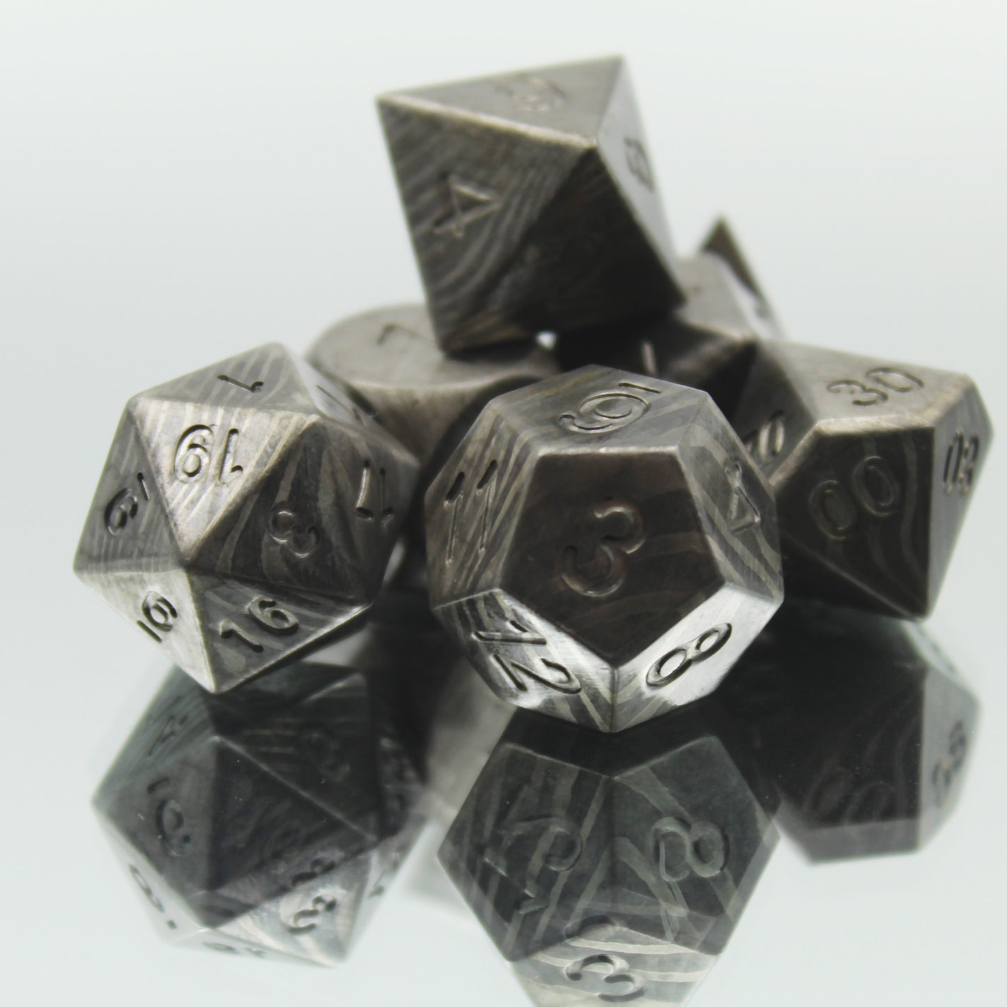 Dice from the Metal Damascus Steel dice set arranged in a pile on a reflective surface with the d20 and d12 dice in front.  These dice have layered banding of light gray on dark gray and no inking on the numbers.