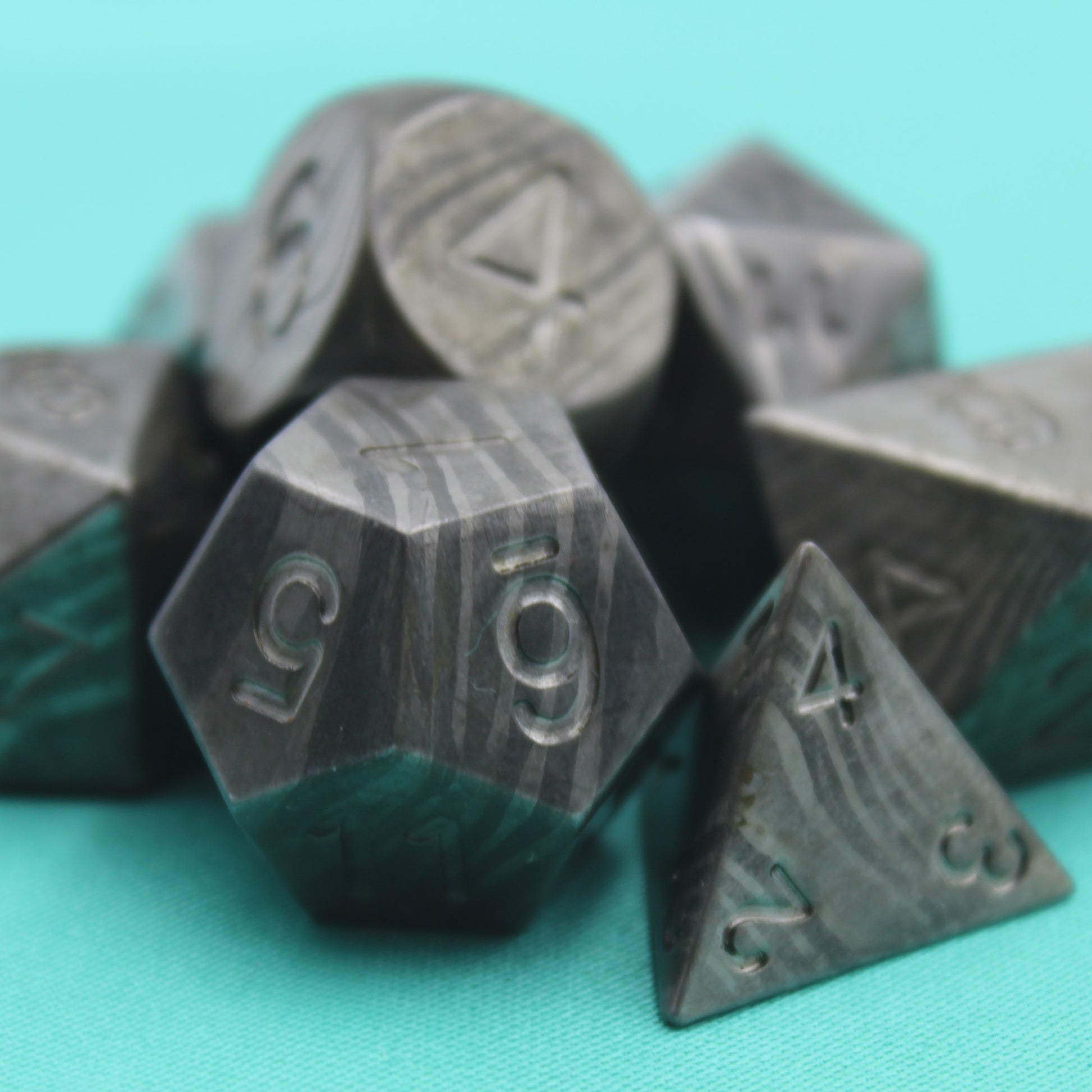 Damascus Steel Dice set arranged in a group on a teal background and surface.  The d12 and d4 are in the foreground showcasing the banding pattern of the dice.