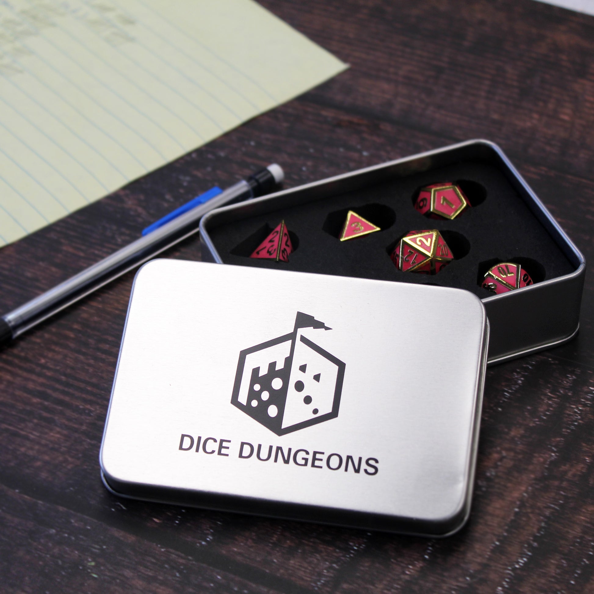Dice Dungeons silver display box on a wooden table, opened partially so that the imperial pink dice inside can be seen. Next to the display box is a mechanical pencil and a piece of yellow lined paper.