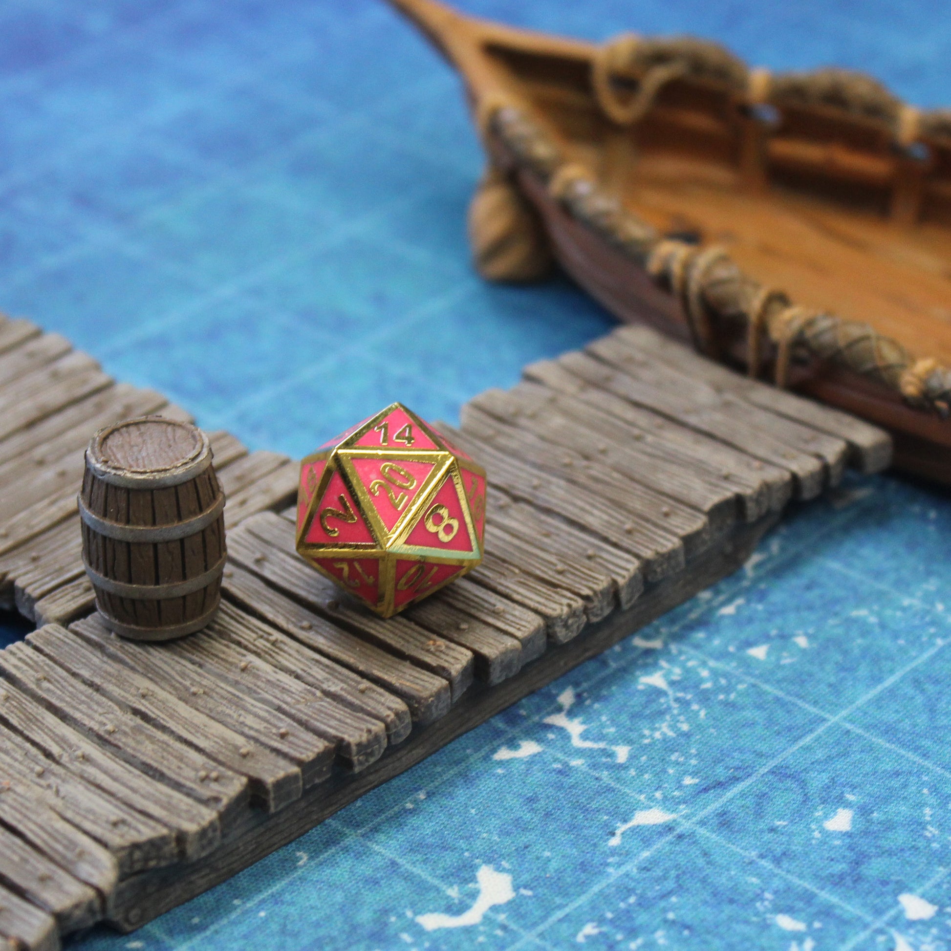 The D20 on a wooden pier set piece on a blue printed map surface. At the end of the pier is a wooden boat, and next to the die is a wooden barrel.
