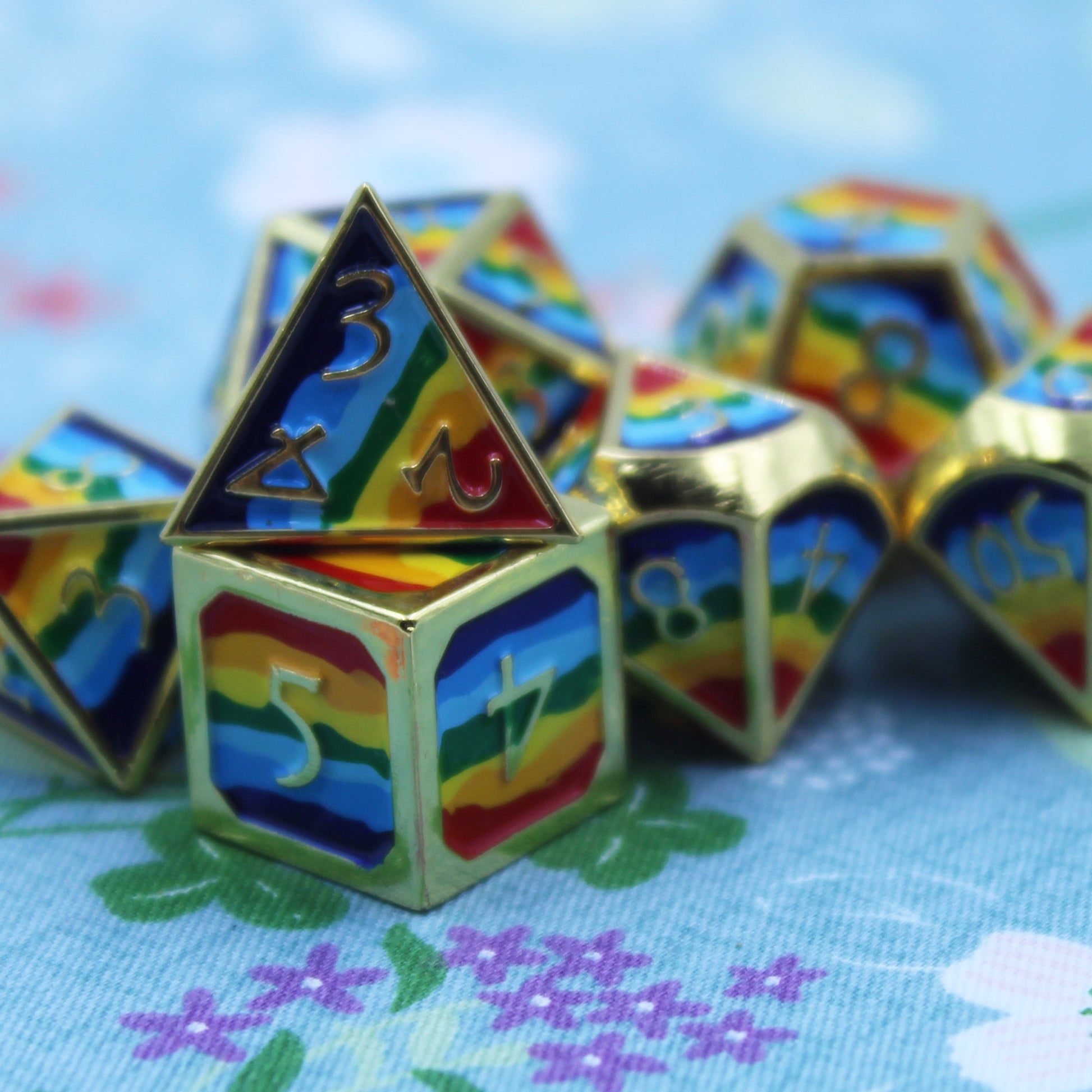 Closeup photograph of rainbow-striped RPG dice with golden numbers and edges on a floral background.
