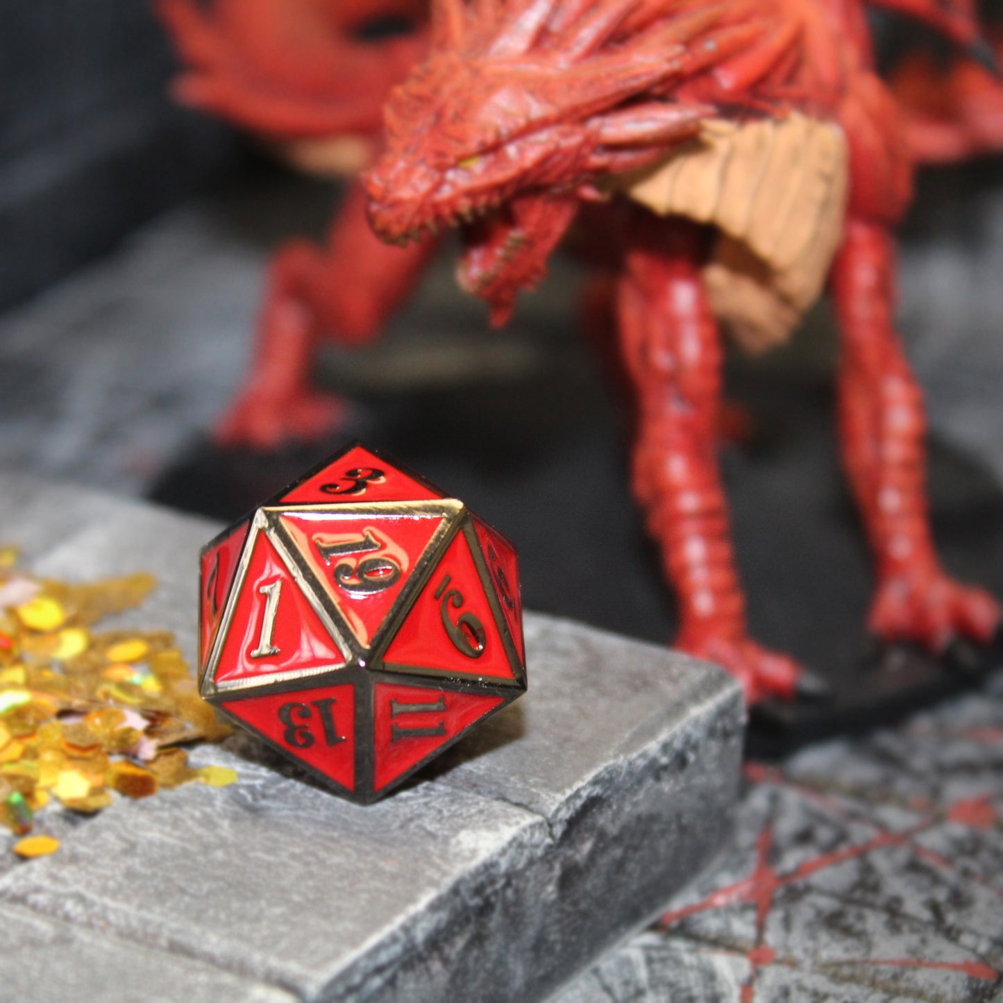 Metal Imperial Red D20 on top of a raised rock set piece, with small gold pieces and a red dragon next to it. The red facing and gold framing on the die are highlighted by the close-up image.