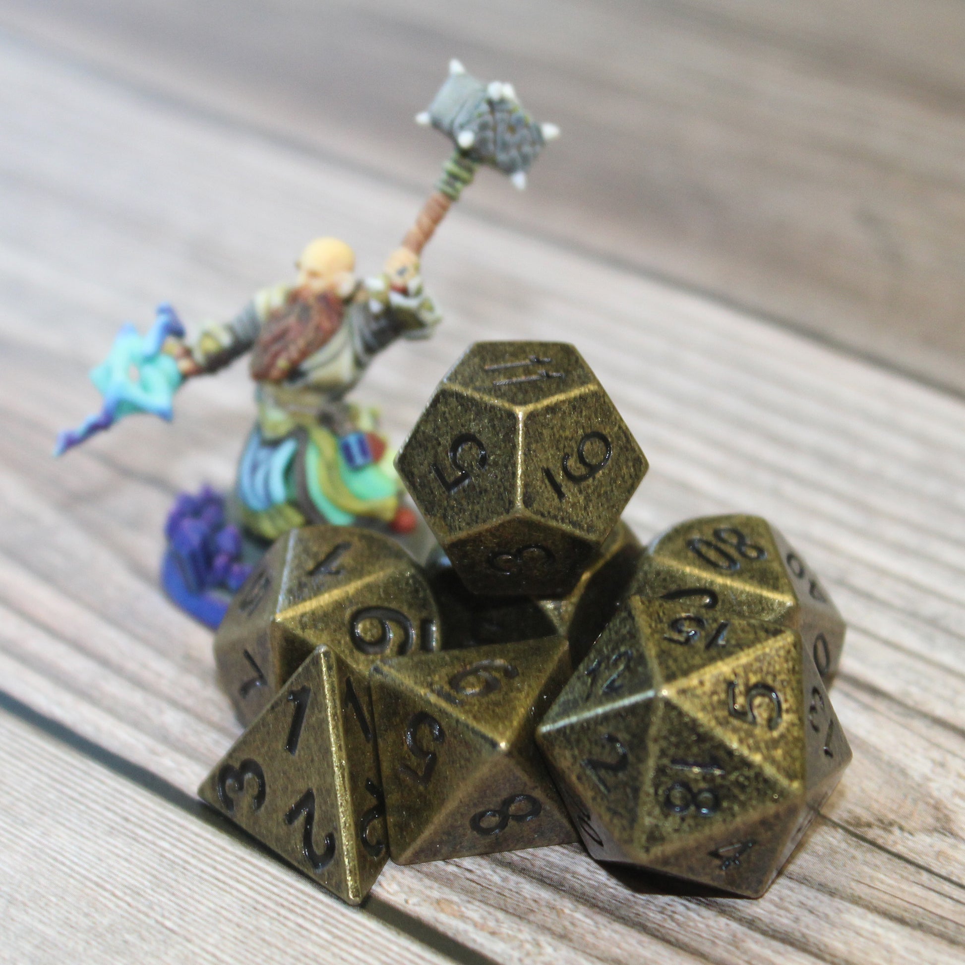 Primordial gold dice set arranged on a light wooden surface with a mini figure next to them for scale
