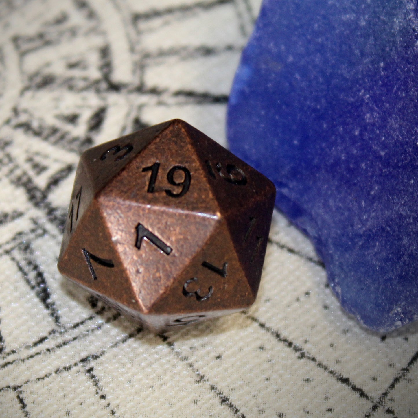 Close-up of Primordial copper D20 die on top of a tan play mat, next to a blue stone.