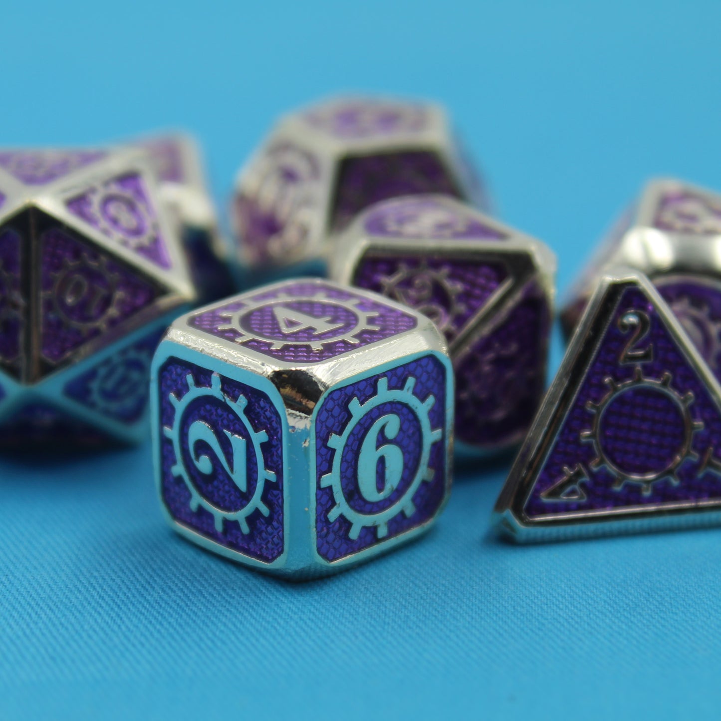 Metal Mechanist dice set arranged in a scattered pile on a blue cloth, with the D6 in focus to showcase the gear design, silver numbers, and purple sides.