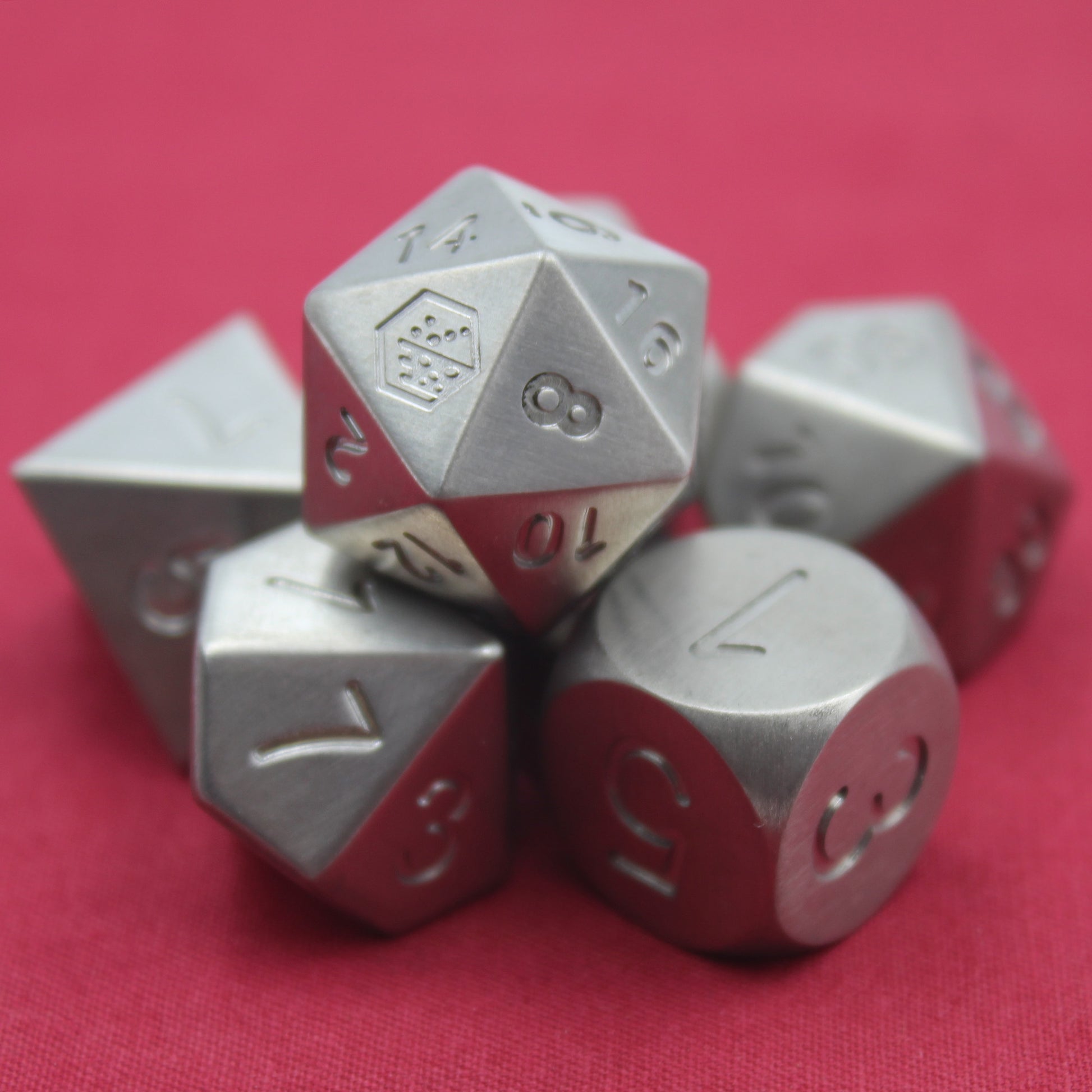 Closeup photo of the Metal Titanium dice set arranged in a pile on a red background with the d20 resting on top with the 8 face towards the viewer and the Dice Dungeons logo in place of the 20 adjacent to the 8 also visible.