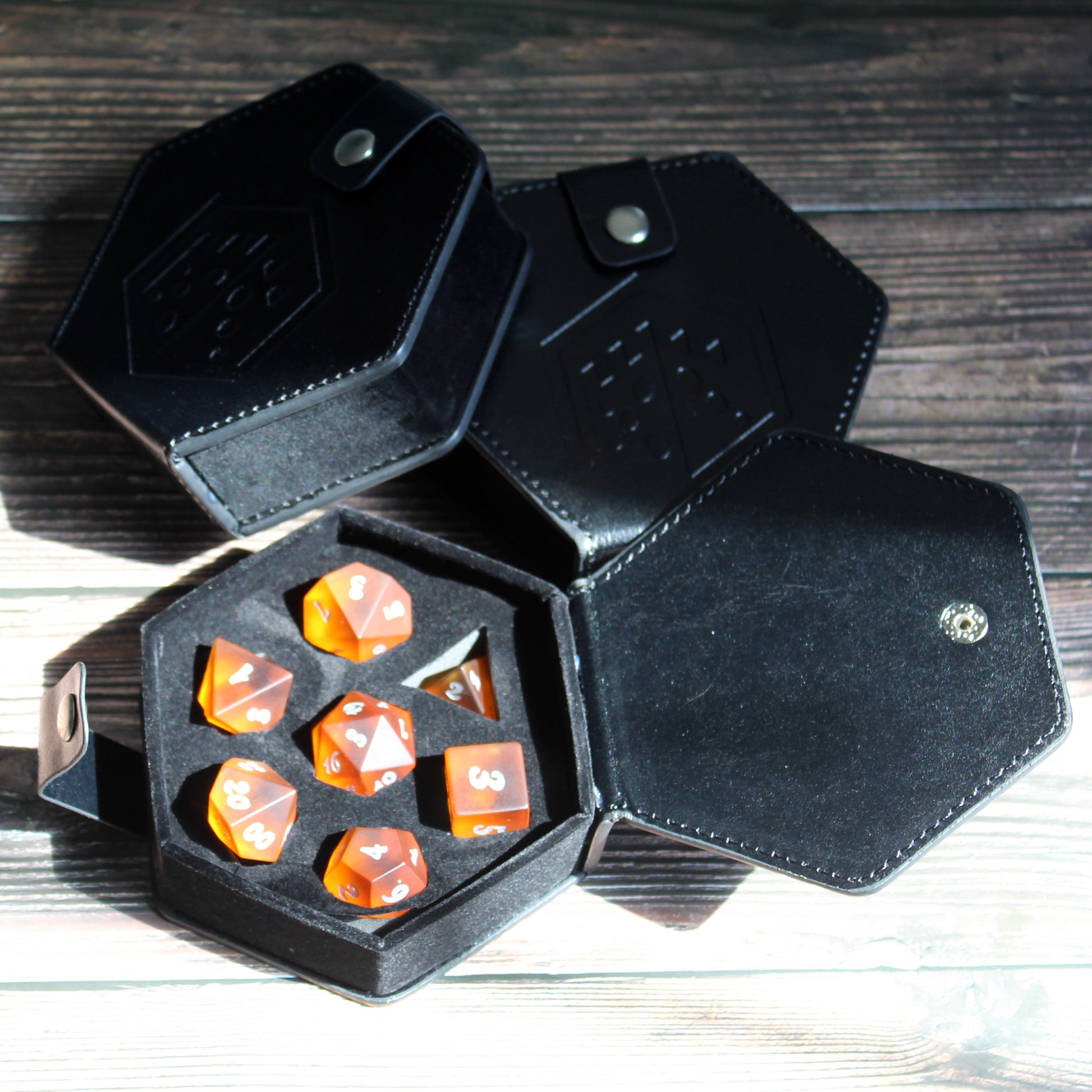 Photograph of a few black faux leather hexagonal dice cases with a silver snap enclosure and the Dice Dungeons logo embossed onto the front of them. One is open and contains a set of orange gaming dice.