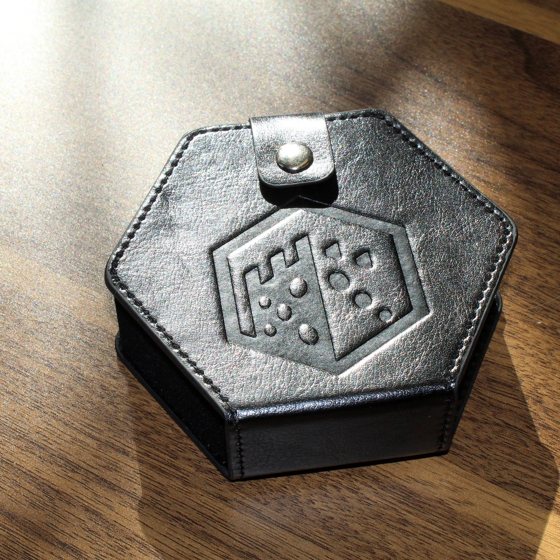A black faux leather hexagonal dice case with a silver snap enclosure and the Dice Dungeons logo embossed onto the front of it on a wooden surface.