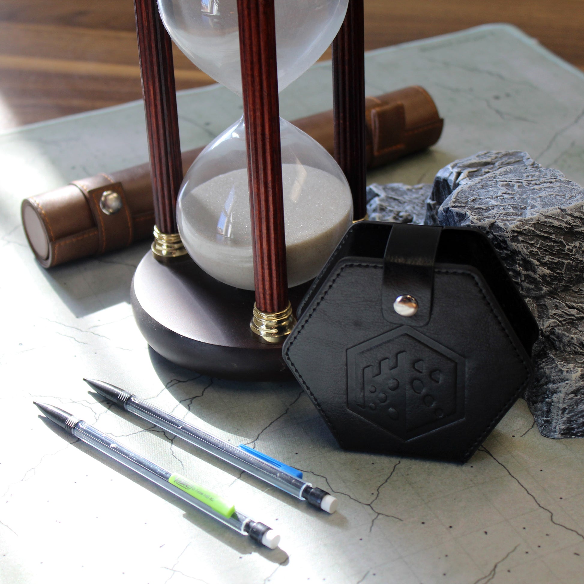 A black faux leather hexagonal dice case with a silver snap enclosure and the Dice Dungeons logo embossed onto the front of it sitting next to an hourglass, some mechanical pencils, and a leatherette scroll case on a gaming mat.