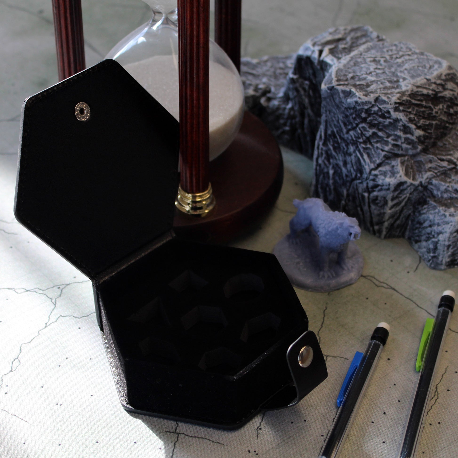 An opened black faux leather hexagonal dice case with a silver snap enclosure sitting amongst gaming supplies on a battle mat.