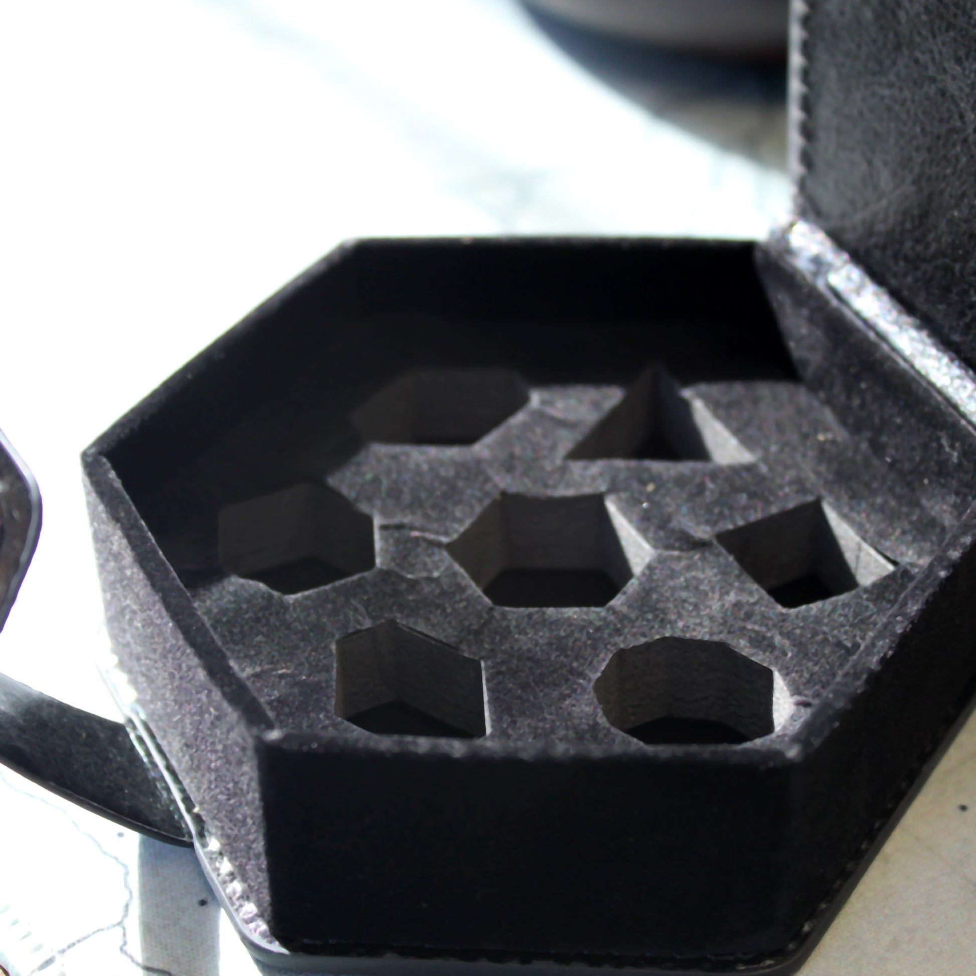 Closeup of an opened An opened black faux leather hexagonal dice case showing the detail of the slots for dice to fit into.