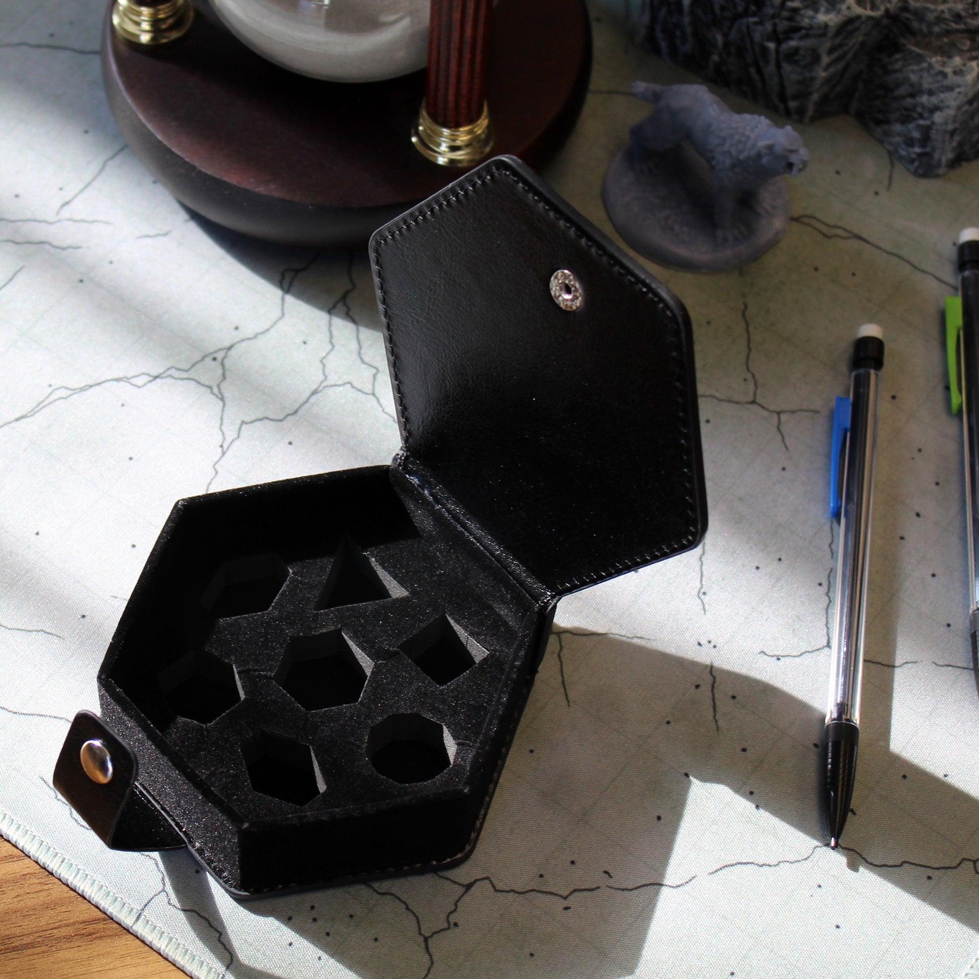 An opened black faux leather hexagonal dice case with a silver snap enclosure sitting amongst gaming supplies on a battle mat.