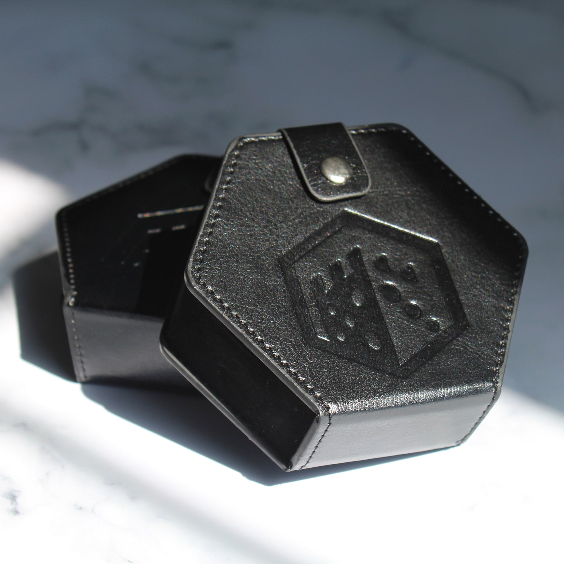 Two black faux leather hexagonal dice cases with a silver snap enclosure and the Dice Dungeons logo embossed onto the front of them leaning against each other on a white marbled surface.