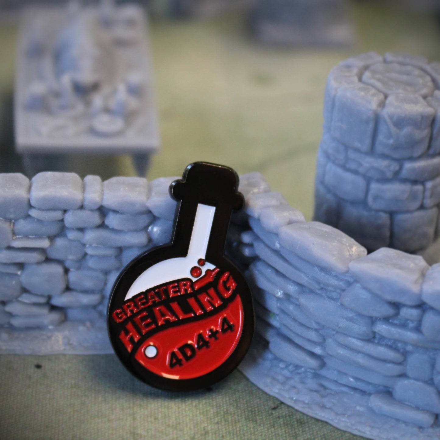 Potion of Greater Healing token is propped up on a gray stone wall set piece on top of a tan game map. Behind the stone wall a grey pillar set piece and a grey cluttered table set piece can be seen.