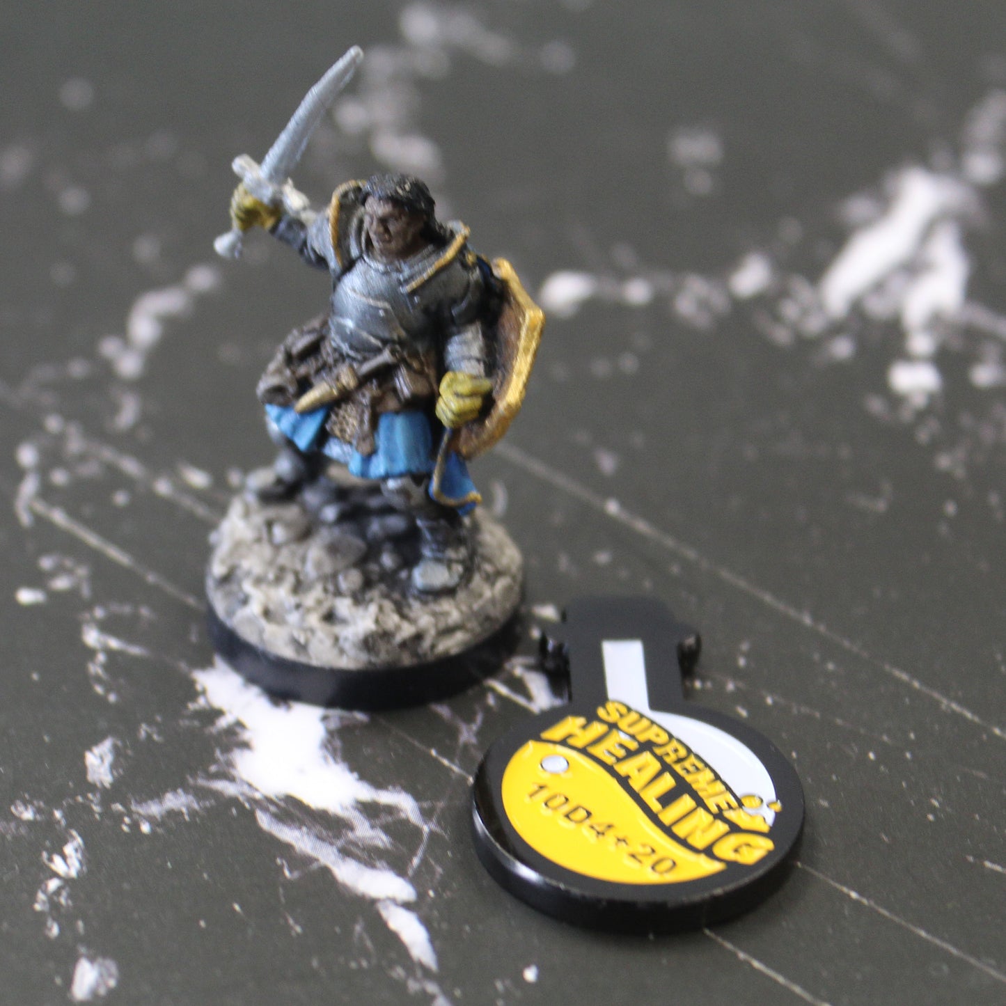 Potion of Supreme Healing token laying on a black surface with white streaking pattern. To the left of the token is a mini figure holding up a sword.