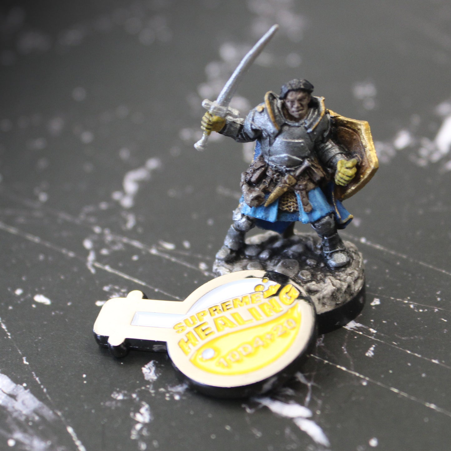 Potion of Supreme Healing Token slightly propped up on a mini figure holding a sword. The way the token is propped up catches the light so that the entire token looks gold. Both the token and the mini figure are on top of a black surface with a white streaking pattern.