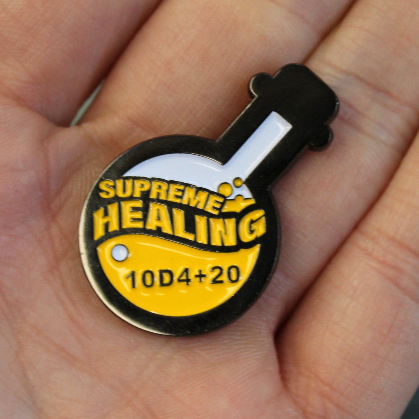 Potion of Supreme Healing Token in the palm of someone's hand. Token is shaped like a round bottom boiling flask, halfway filled with the design of yellow bubbly liquid and the words "supreme healing" written in yellow. On the liquid is the black lettering "10D4+20".