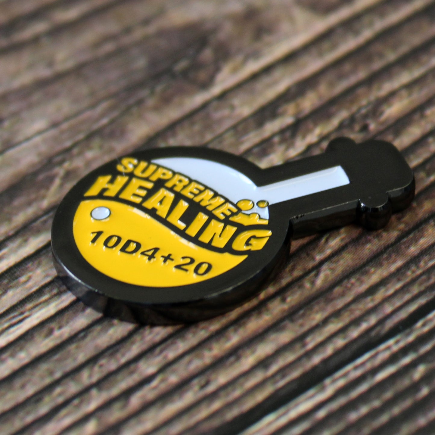 Potion of Supreme Healing Token on dark wood surface. Token is shaped like a round bottom boiling flask, halfway filled with the design of yellow bubbly liquid and the words "supreme healing" written in yellow. On the liquid is the black lettering "10D4+20".