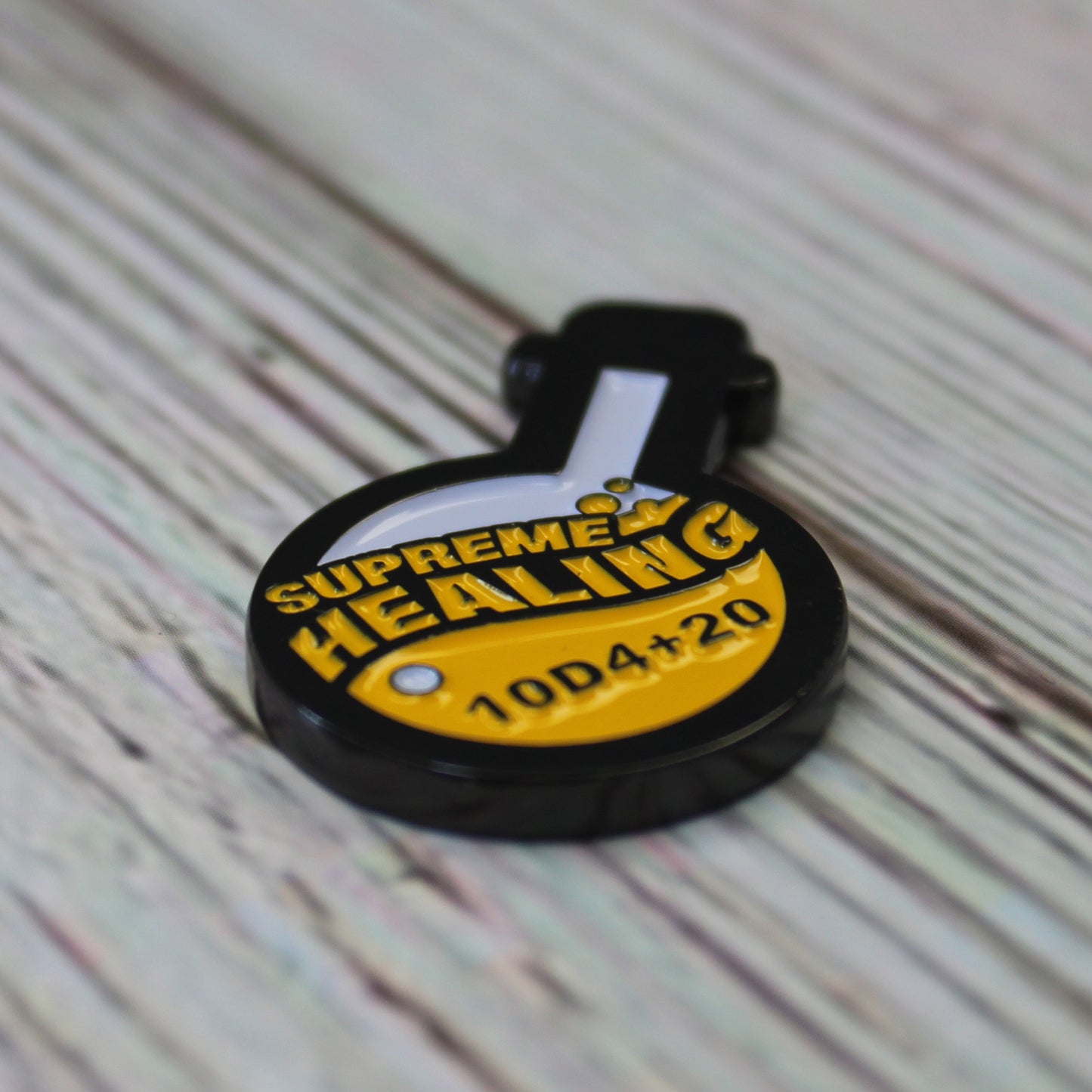 Potion of Supreme Healing Token on white wood surface. Token is shaped like a round bottom boiling flask, halfway filled with the design of yellow bubbly liquid and the words "supreme healing" written in yellow. On the liquid is the black lettering "10D4+20".