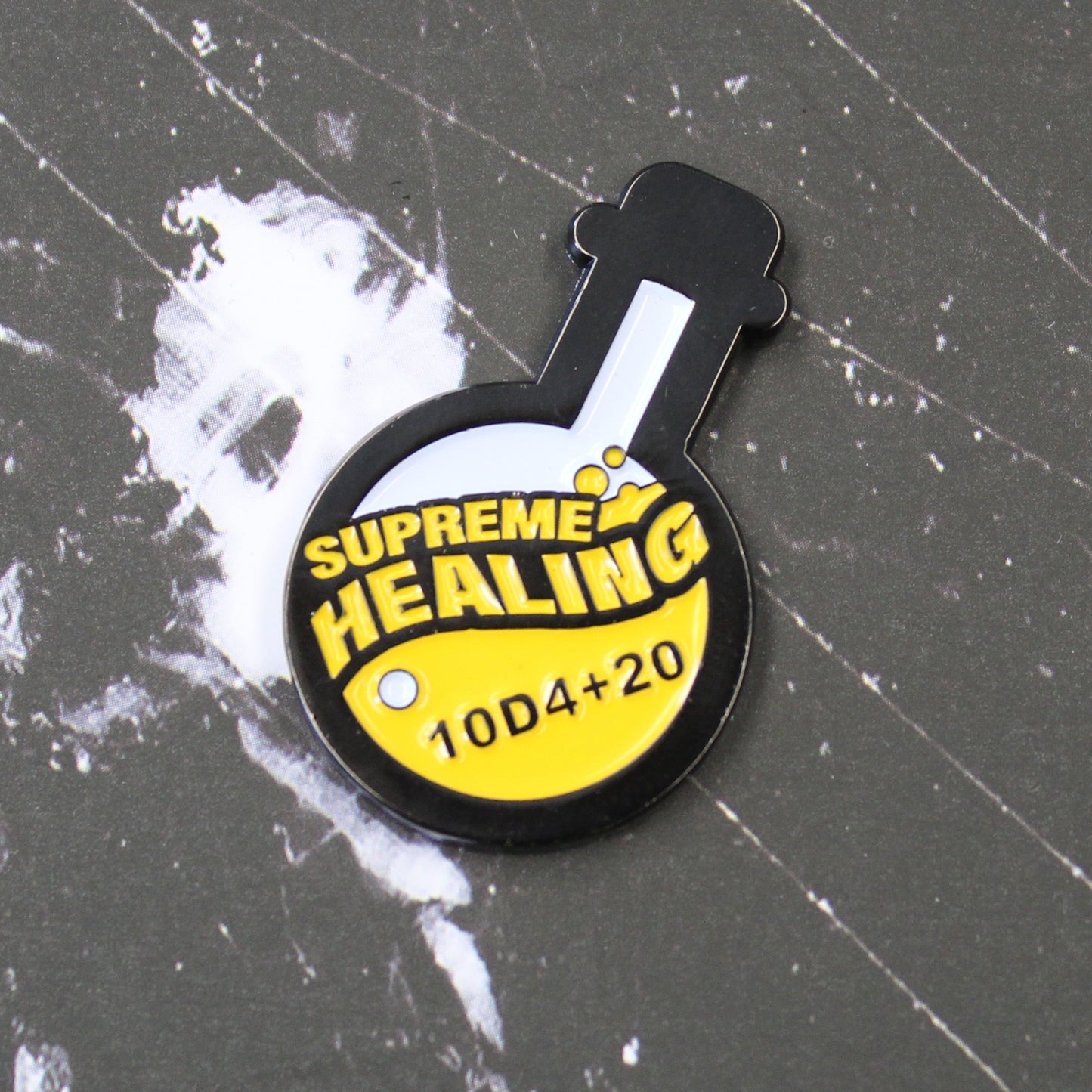 Potion of Supreme Healing Token on black surface with white streaking pattern. Token is shaped like a round bottom boiling flask, halfway filled with the design of yellow bubbly liquid and the words "supreme healing" written in yellow. On the liquid is the black lettering "10D4+20".
