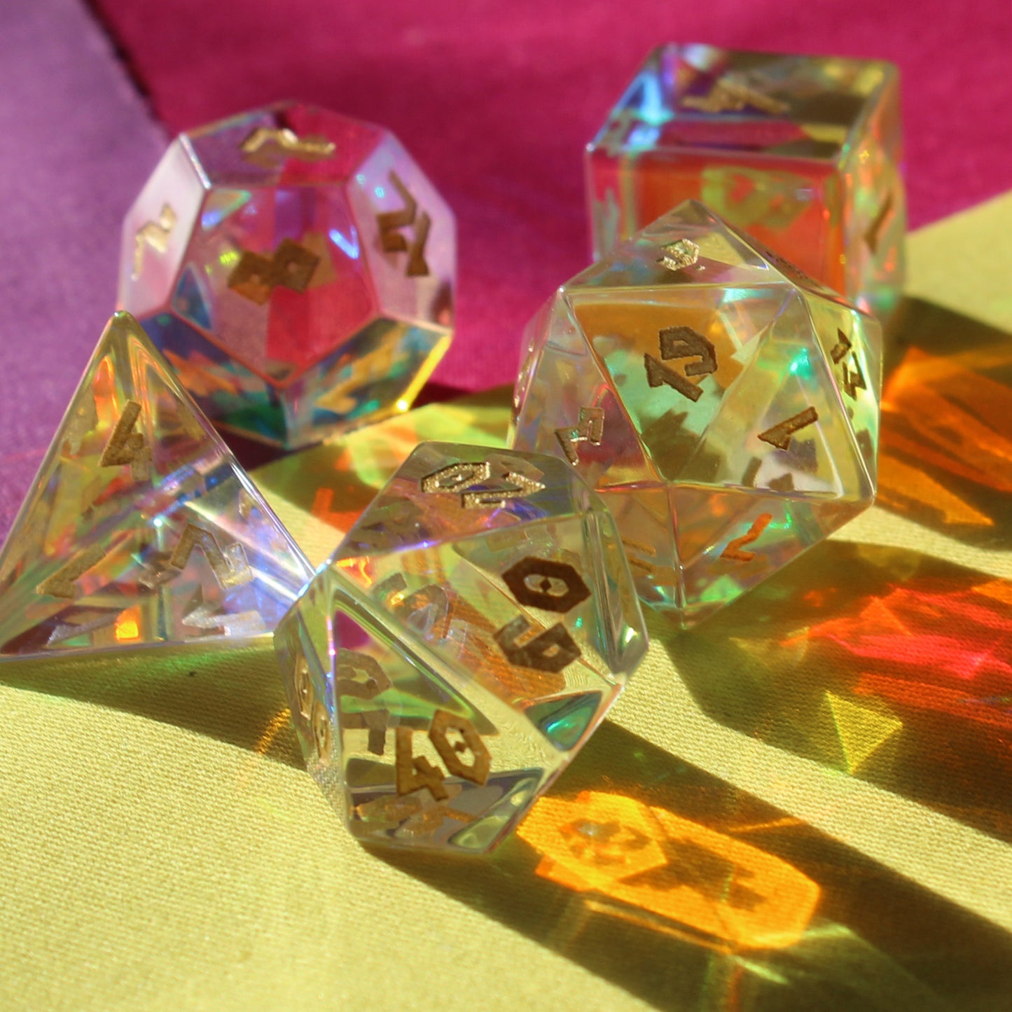 Close up shot of Prismatic Glass dice refracting sunlight on a multi colored cloth surface. The 19 face of the d20 faces the viewer and the detail of the etched numbering is clearly visible on the d10 in the foreground.