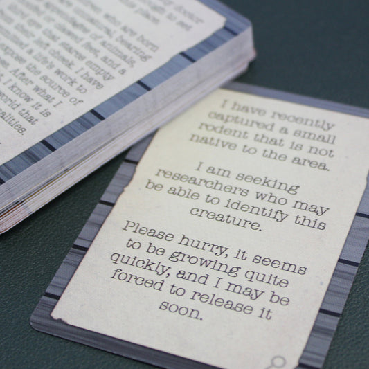 A close up of one card next to a stack of other cards from the deck on a black table. The cards have a gray wood pattern around the edges with white in the center and black lettering. The card says "I have recently captured a small rodent that is not native to the area. I am seeking researchers who may be able to identify this creature. Please hurry, it seems to be growing quite quickly, and I may be forced to release it soon."