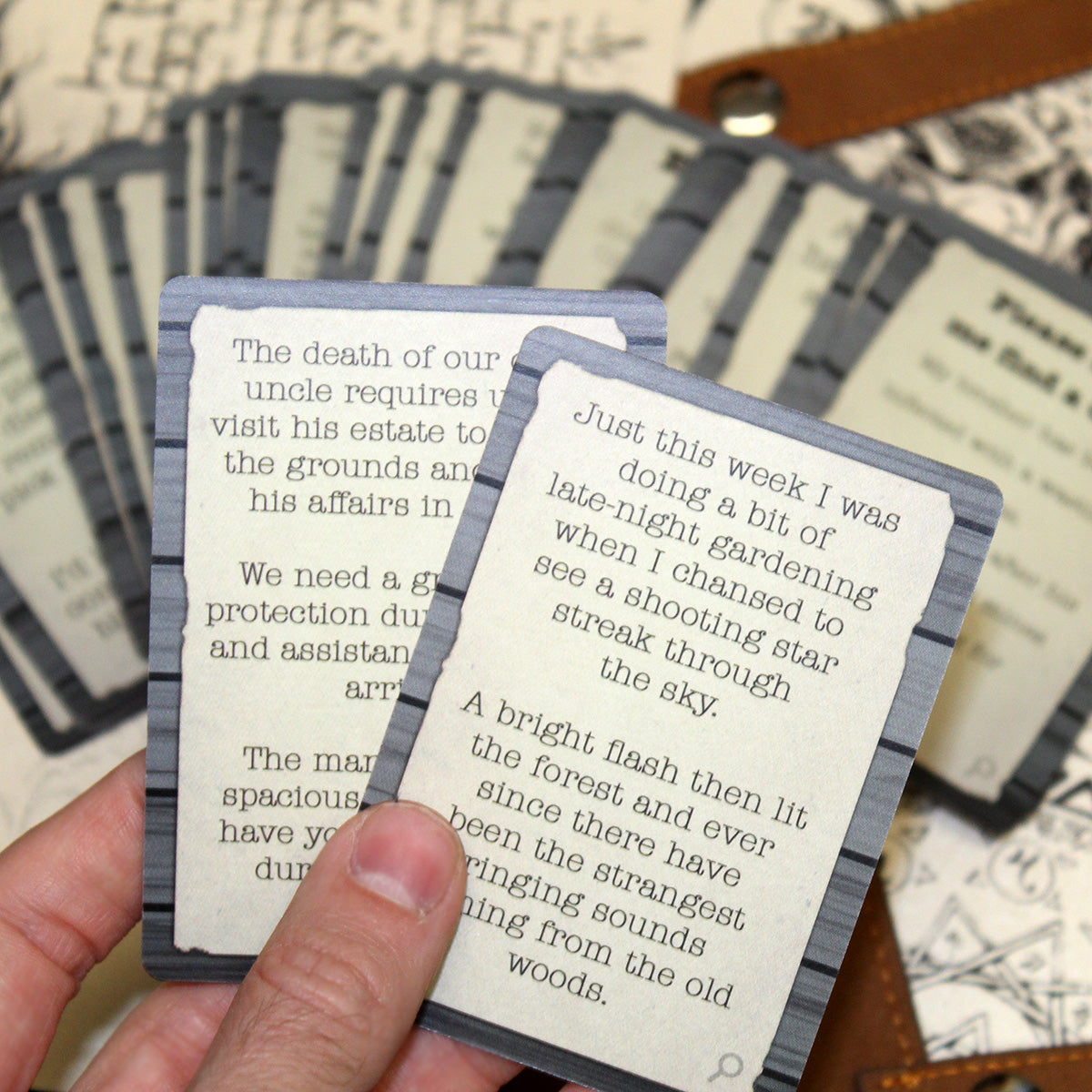 Two cards are being held in someone's hand above a table. The card on top says "Just this week I was doing a bit of late-night gardening when I chansed to see a shooting star streak through the sky. A bright flash then lit the forest and ever since there have been the strangest ringing sounds coming from the old woods." On the table the rest of the cards are fanned out on top of a white cloth with an intricate black design.