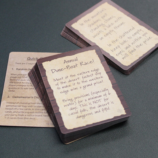 The card from the deck are divided into two piles and laid out on a gray surface as if they are in mid-use. The cards are tan with fake wood grain around the edges and brown lettering. The card in focus says "Annual Dune-Boat Race!" and describes scenario on the rest of the card.