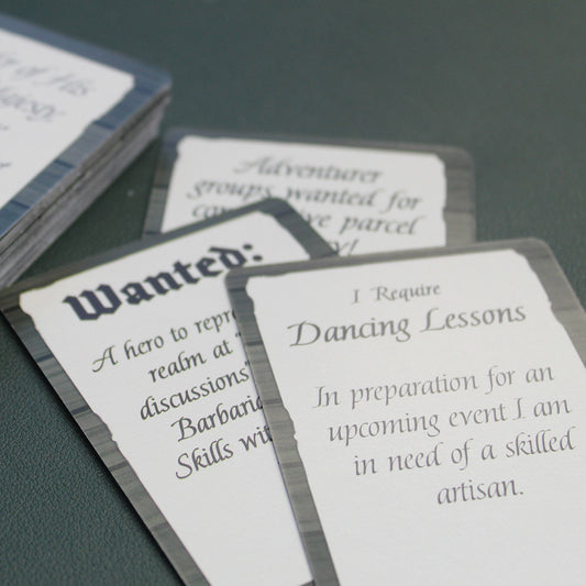Closeup of three of the cards from the deck laid down as if in mid-use on a black table. The card on top reads "I require dancing lessons in preparation for an upcoming event I am in need of a skilled artisan." All that can be read on the card underneath it is  "Wanted".