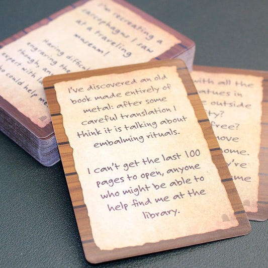 Two small stacks of cards from the deck are perpendicular to each other, and one stack is placed slightly on top of the other, on top of a black table. The stack in focus has a card on top that says "I've discovered an old book made entirely of metal: after some careful translation I think it is talking about embalming rituals. I can't get the last 100 pages to open, anyone who might be able to help find me at the library."