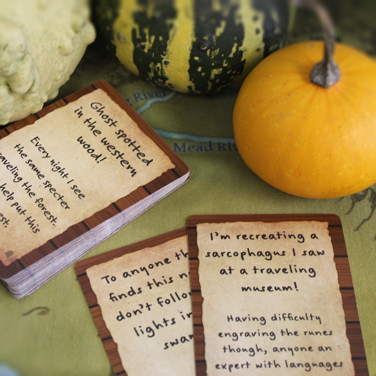 Two small stacks of cards from the deck are perpendicular to each other on top of a green game map. The cards say things like "Ghost spotted in the western wood!" and "I'm recreating a sarcophagus I saw at a traveling museum!" Behind the cards are three small gourds, white, green and yellow striped, and orange.