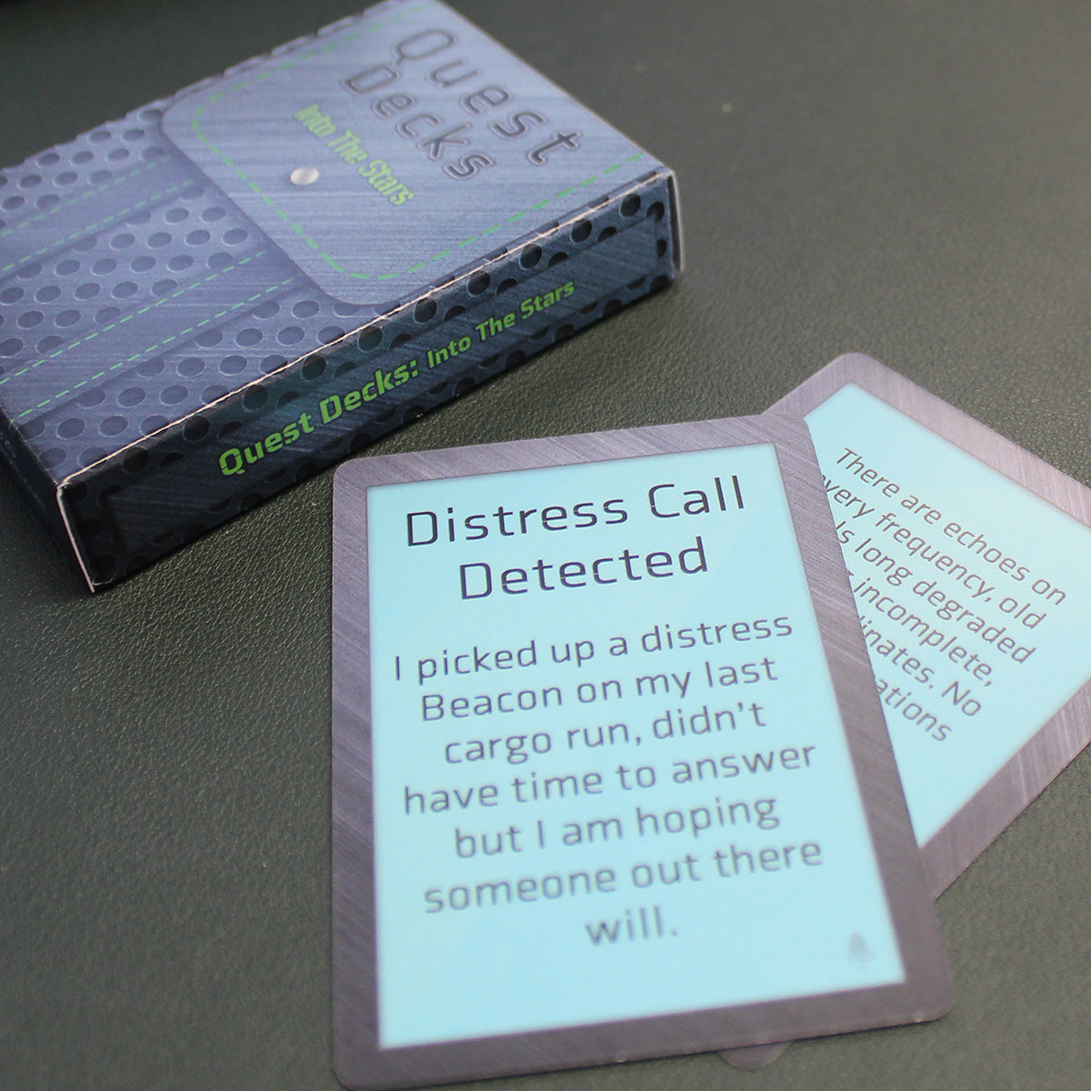 The Into the Stars Quest Deck box is laying on a black table with two cards laying next to it. The card on top reads "Distress Call Detected I picked up a distress Beacon on my last cargo run, didn't have time to answer but I am hoping someone out there will."