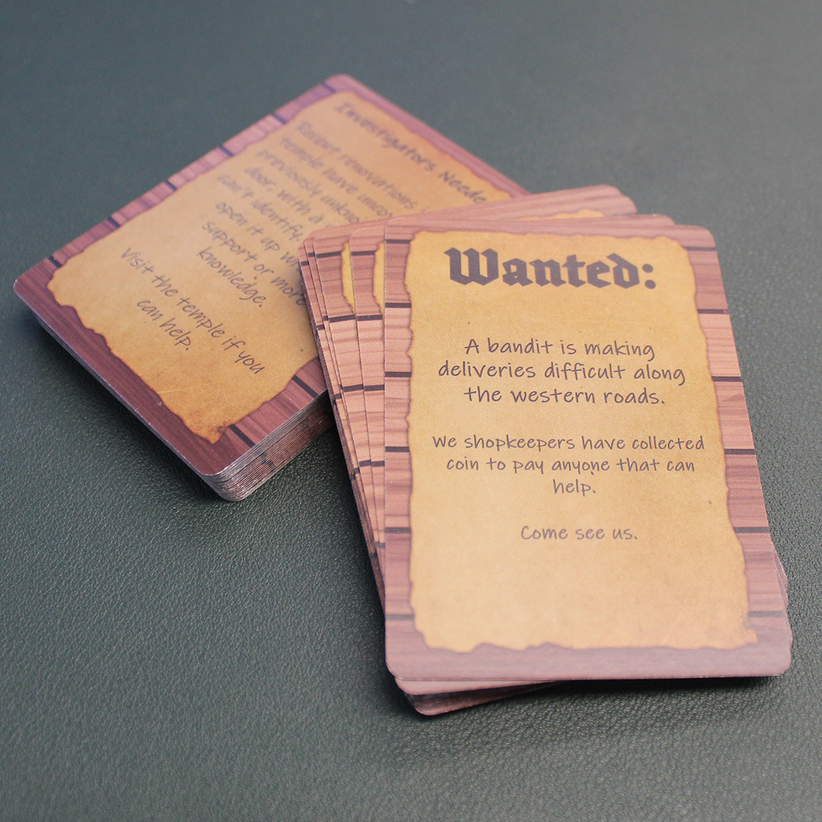 The deck has been split into two stacks, with the stack on top propped up slightly on the lower stack on a black table. The card on the top of the stack reads "Wanted: A bandit is making deliveries difficult along the western roads. We shopkeepers have collected coin to pay anyone that can help. Come see us."