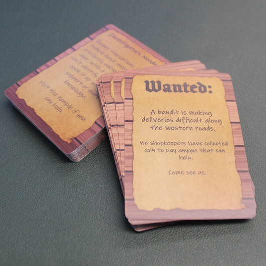 The deck has been split into two stacks, with the stack on top propped up slightly on the lower stack on a black table. The card on the top of the stack reads "Wanted: A bandit is making deliveries difficult along the western roads. We shopkeepers have collected coin to pay anyone that can help. Come see us."
