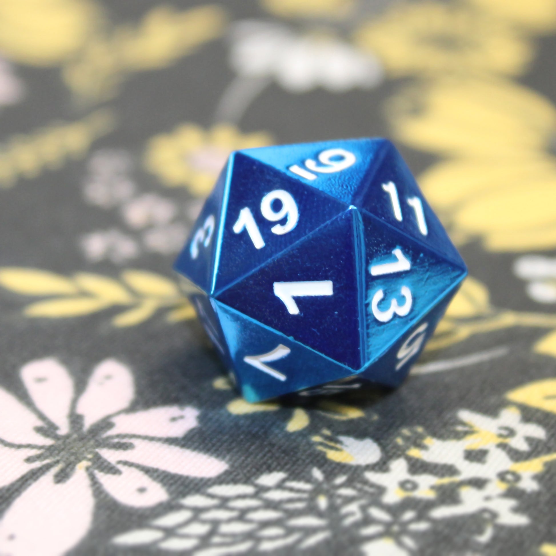 Close-up of the Radiant blue D20 on a black floral fabric with pink, white, and yellow flowers. The close-up showcases the metallic blue of the faces and the white numbering.