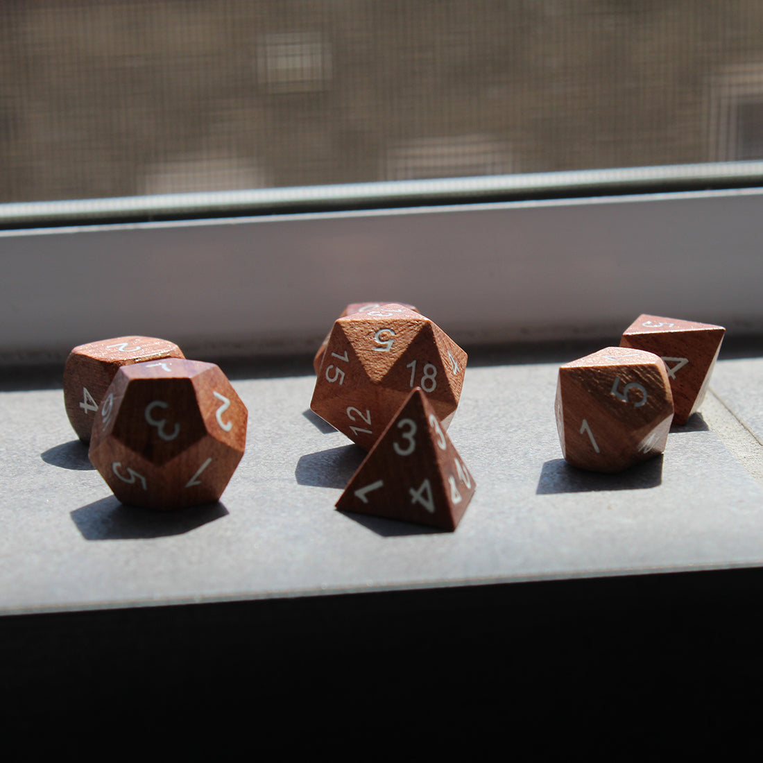 A picture of Rose Wood Wooden dice set on a slate windowsill in sunlight.  Each die is carved from solid rosewood with white inked numbering for readability.