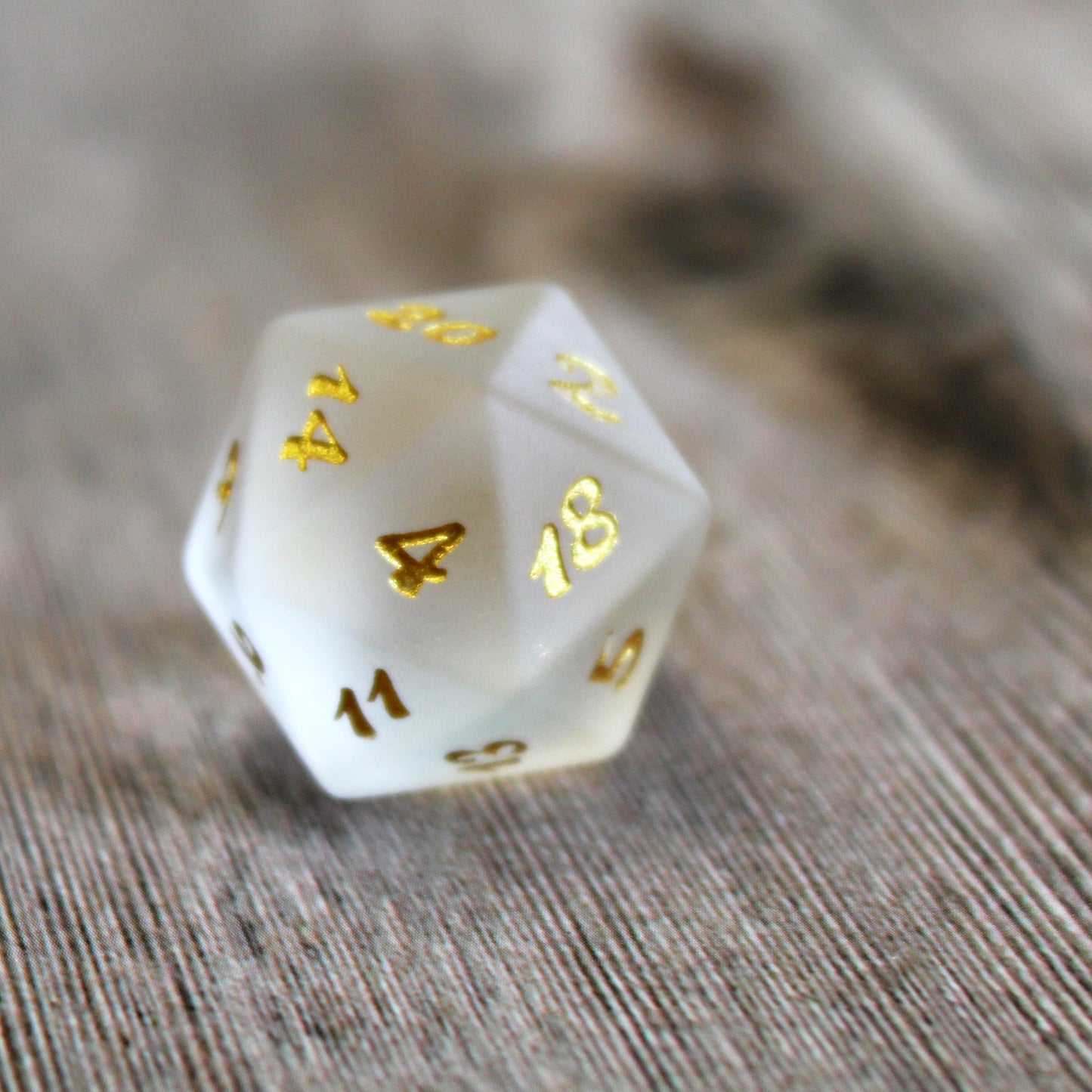 A closeup of a single frosted white glass twenty-sided die with gold numbering on a wooden background