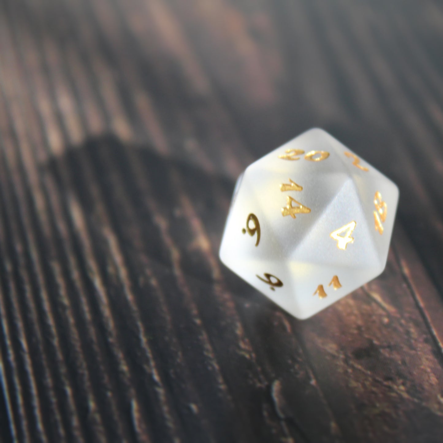 A white frosted glass twenty-sided die with golden numbering on a wooden background