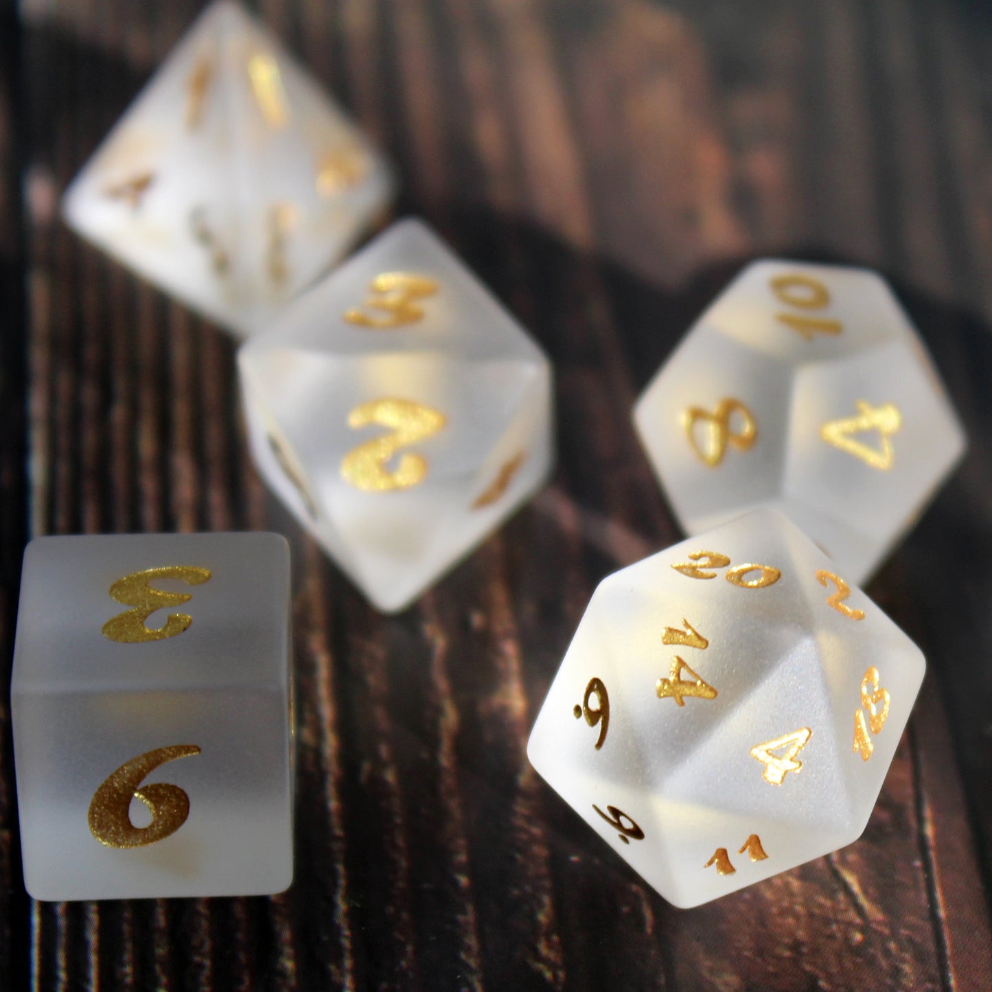 A closeup of part of a set of white frosted glass RPG dice with golden font on a wooden background