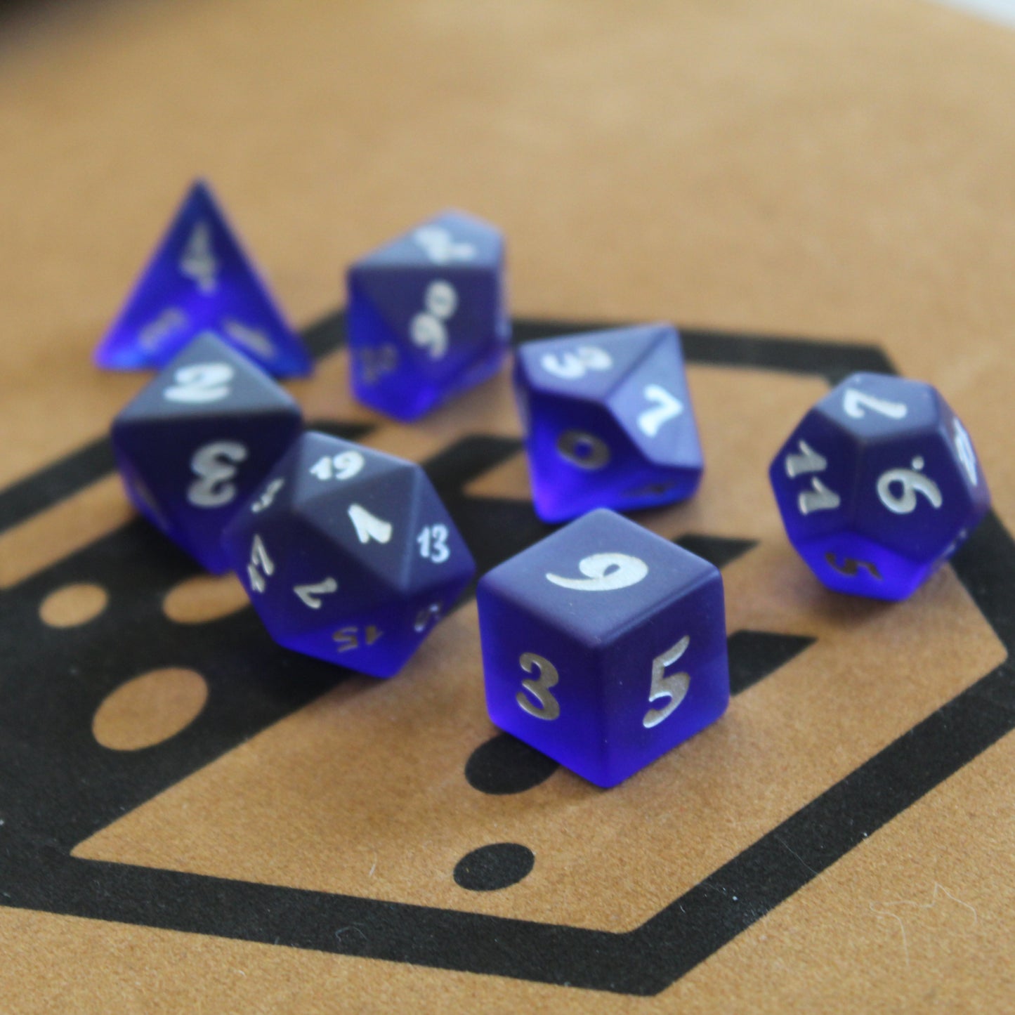 A set of dark blue translucent glass RPG dice with silver numbering on a brown surface displaying the Dice Dungeons logo