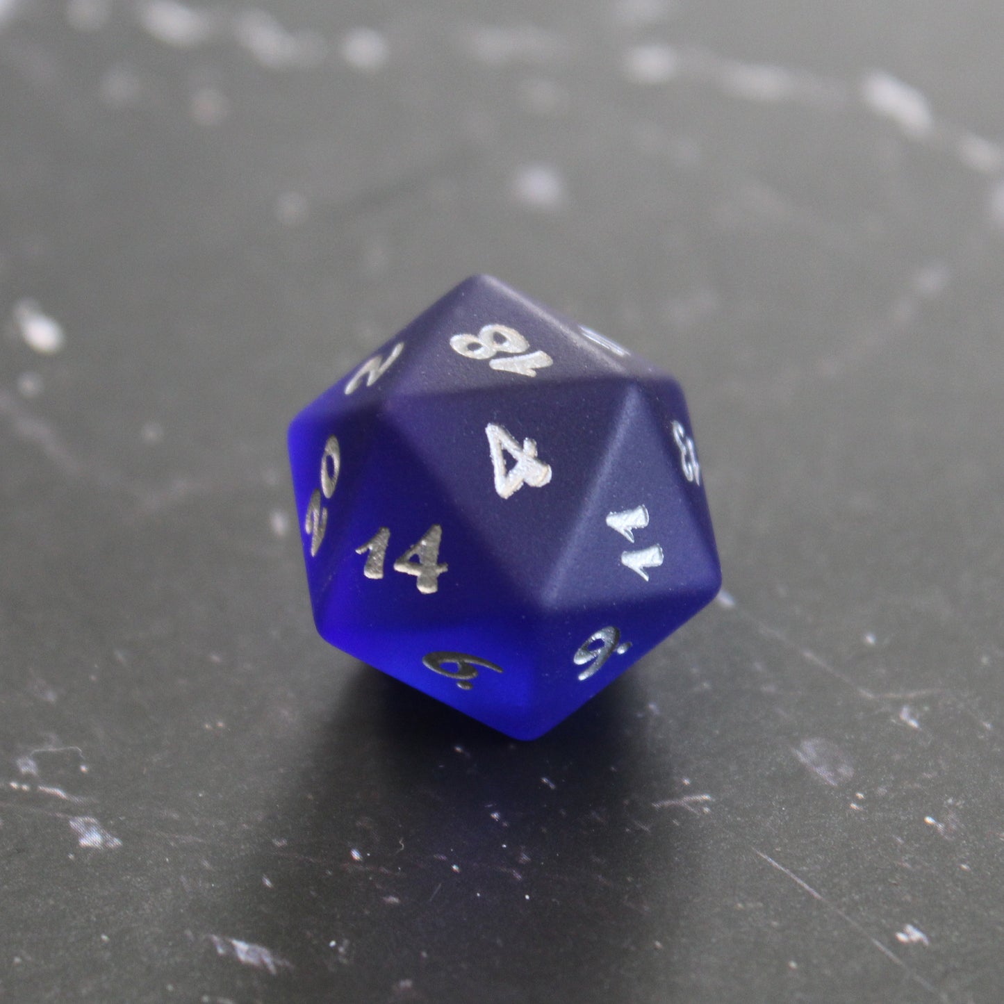 A dark blue translucent glass twenty-sided die with silver numbering on a black marbled surface