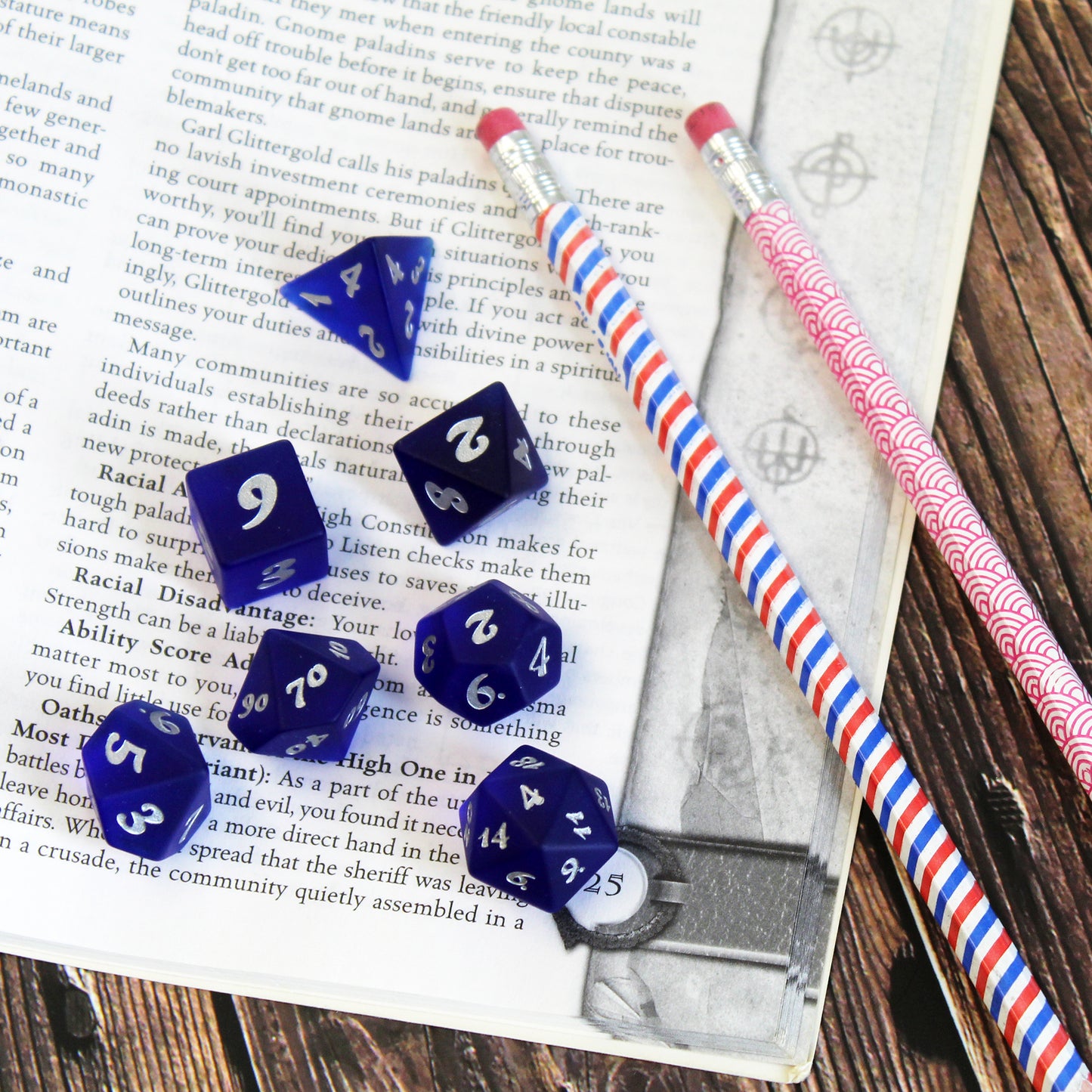 A set of dark blue translucent glass RPG dice with silver numbering on the page of an open book next to some pencils.